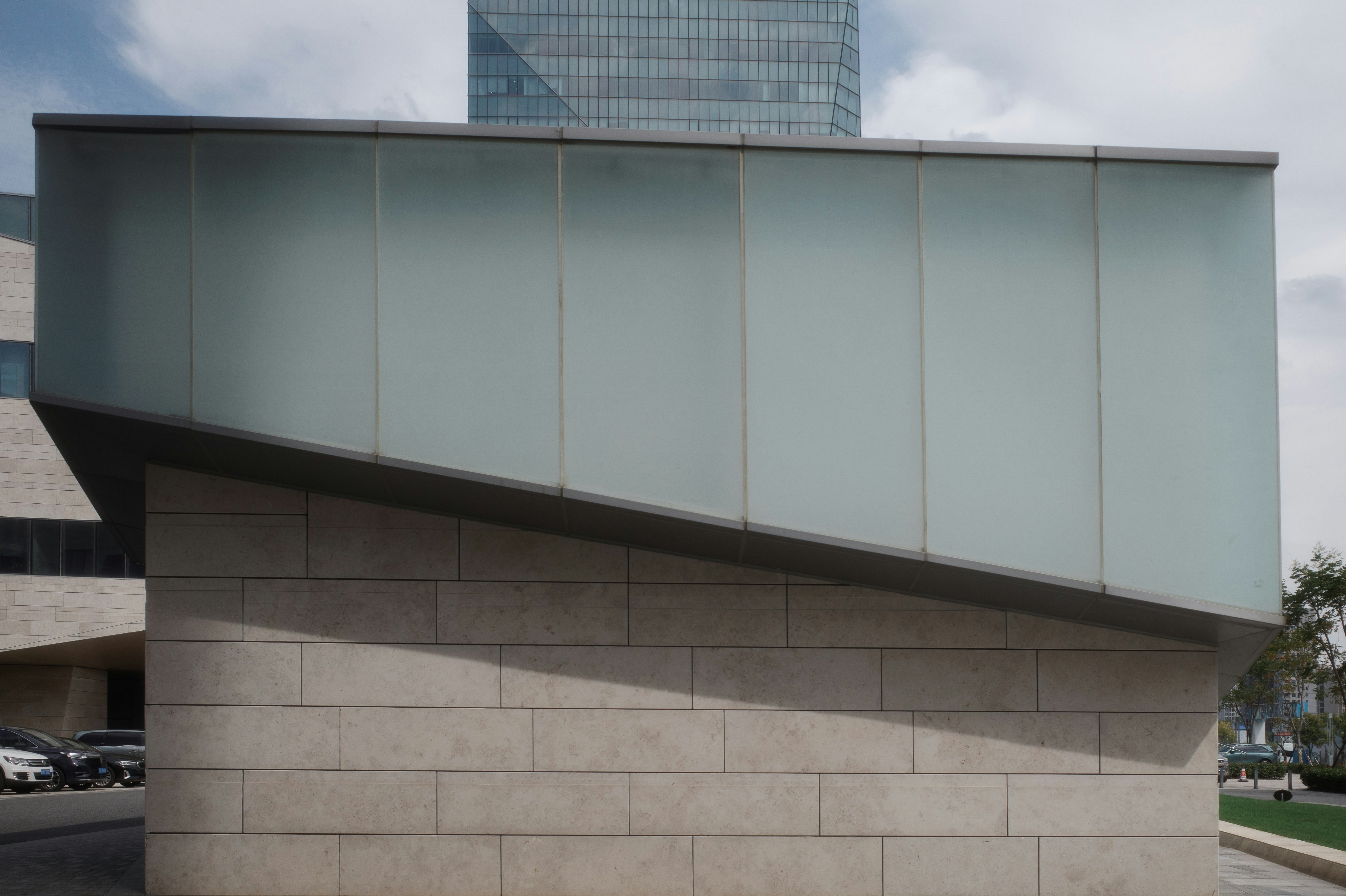 Modern architectural facade featuring an angled frosted glass overhang above a stone wall, set against a cloudy sky.