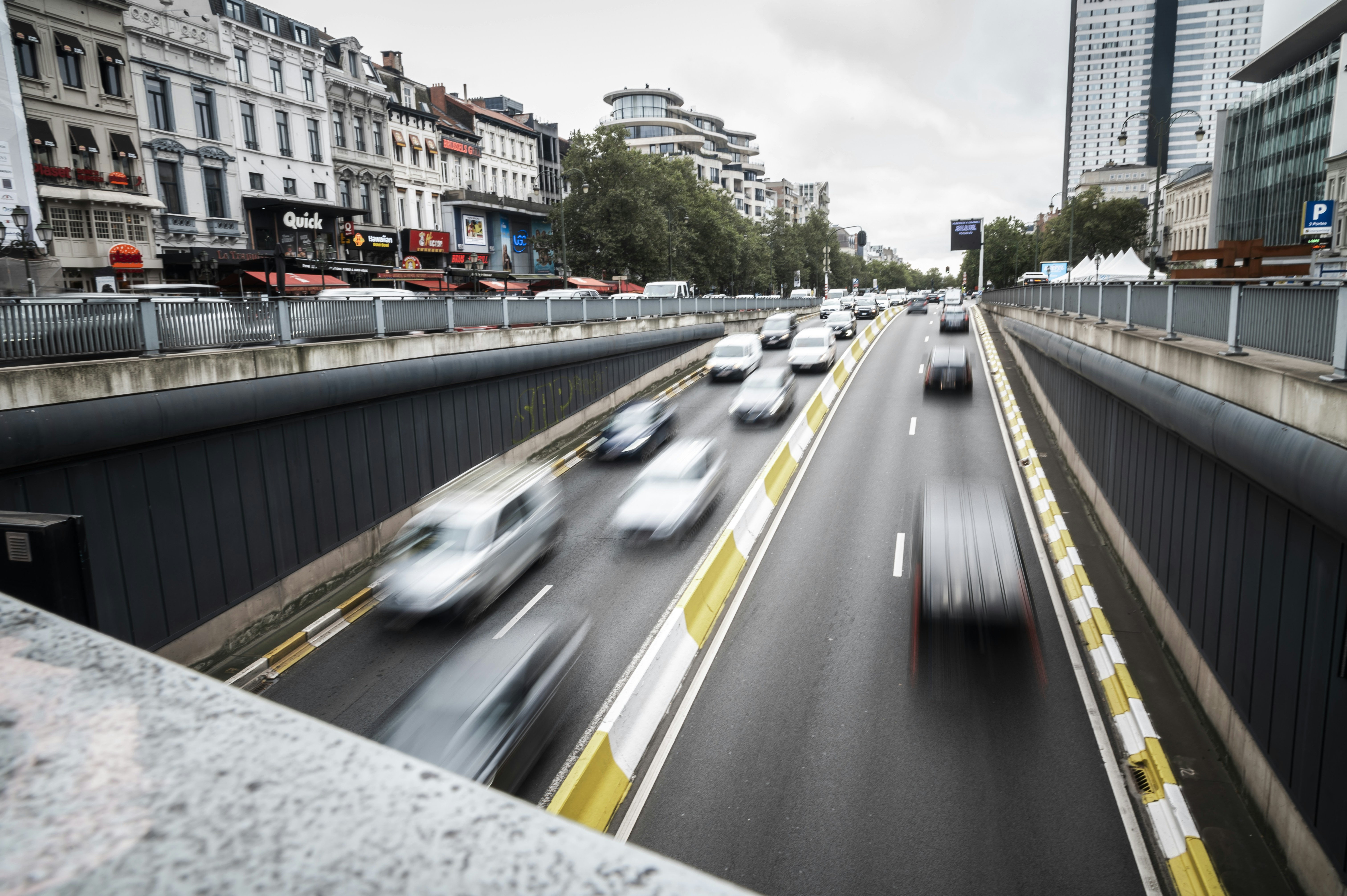Voitures floues entrant dans un tunnel d’un grand boulevard bruxellois, sous un ciel gris