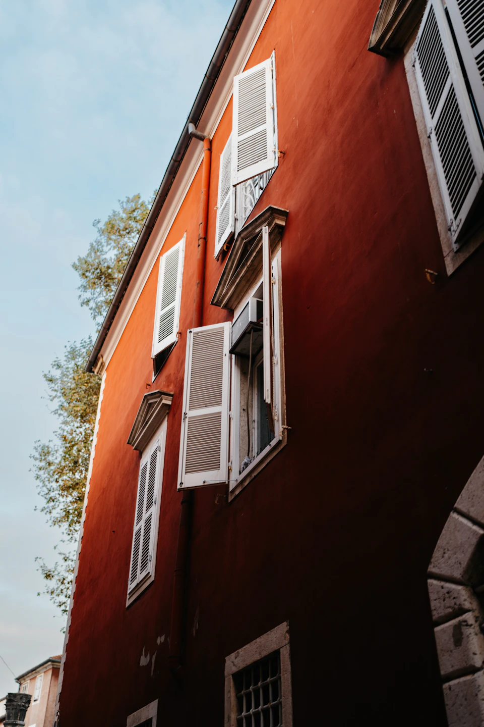 The image shows a red building with white shutters on its windows, set against a clear blue sky. The perspective is from below, looking up at the building's facade and the open windows. The building has six windows, each adorned with white shutters that are slightly ajar, allowing a glimpse of the world outside to enter the room.