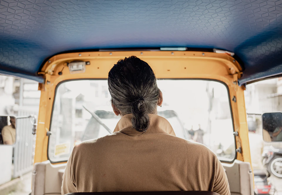 The image shows a person sitting inside an orange and yellow auto rickshaw, facing away from the camera with their back to it. The interior of the vehicle is visible through the window, which has wipers that are currently in use. The car appears to be parked on a street or road, as there are no other vehicles or people visible in the image.