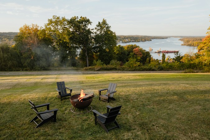 Wood fire burning in a round metal fire pit on a mown lawn, four dark Adirondack chairs around it and a wide river below the trees