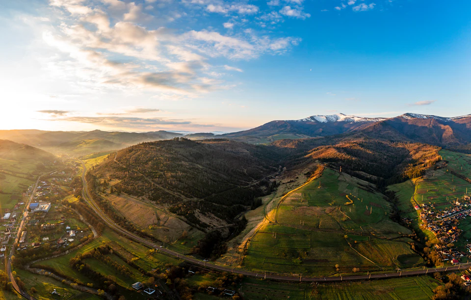 The image presents an aerial view of a valley nestled between two mountains, with a road cutting through it. The valley is lush and green, dotted with trees that add depth to the scene. In the distance, snow-capped mountains rise against the sky, their peaks touching the horizon. The sun casts long shadows across the landscape, creating a beautiful interplay of light and shadow on the terrain below.