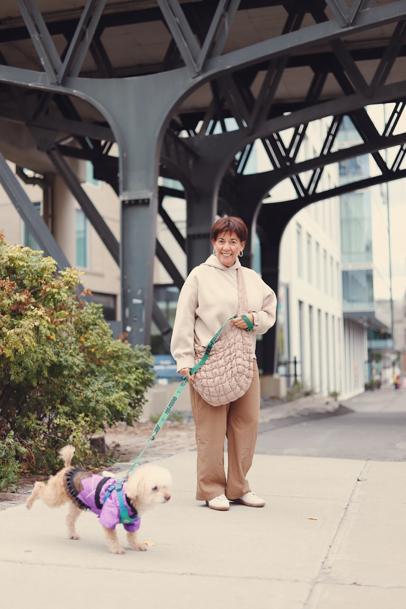 A smiling woman walking a small dog in a coat on a city sidewalk on a calm day