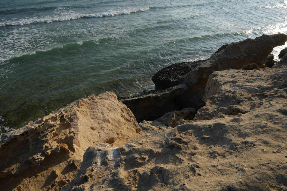 The image depicts a rocky cliff overlooking the ocean, with the vast expanse of water stretching out to meet the sky at the horizon. The rocks on the cliff are jagged and uneven, adding texture to the scene. The colors in the image are predominantly green from the surrounding vegetation and brown from the rocky cliffs. The perspective is from below, looking up towards the cliff, creating a sense of grandeur and scale.