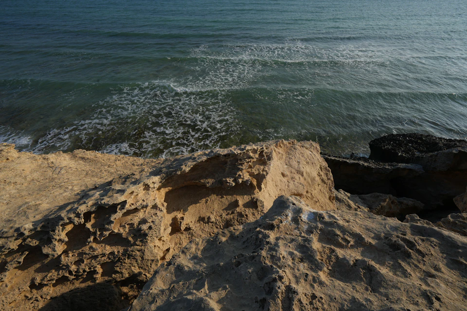 The image depicts a rocky cliff overlooking the ocean, with the vast expanse of water stretching out to meet the sky at the horizon. The rocks on the cliff are jagged and uneven, adding texture to the scene. The colors in the image are predominantly browns and greens, reflecting the natural hues of the landscape. The perspective is from a low angle, looking up towards the ocean, which enhances the grandeur of both the cliff and the sea.
