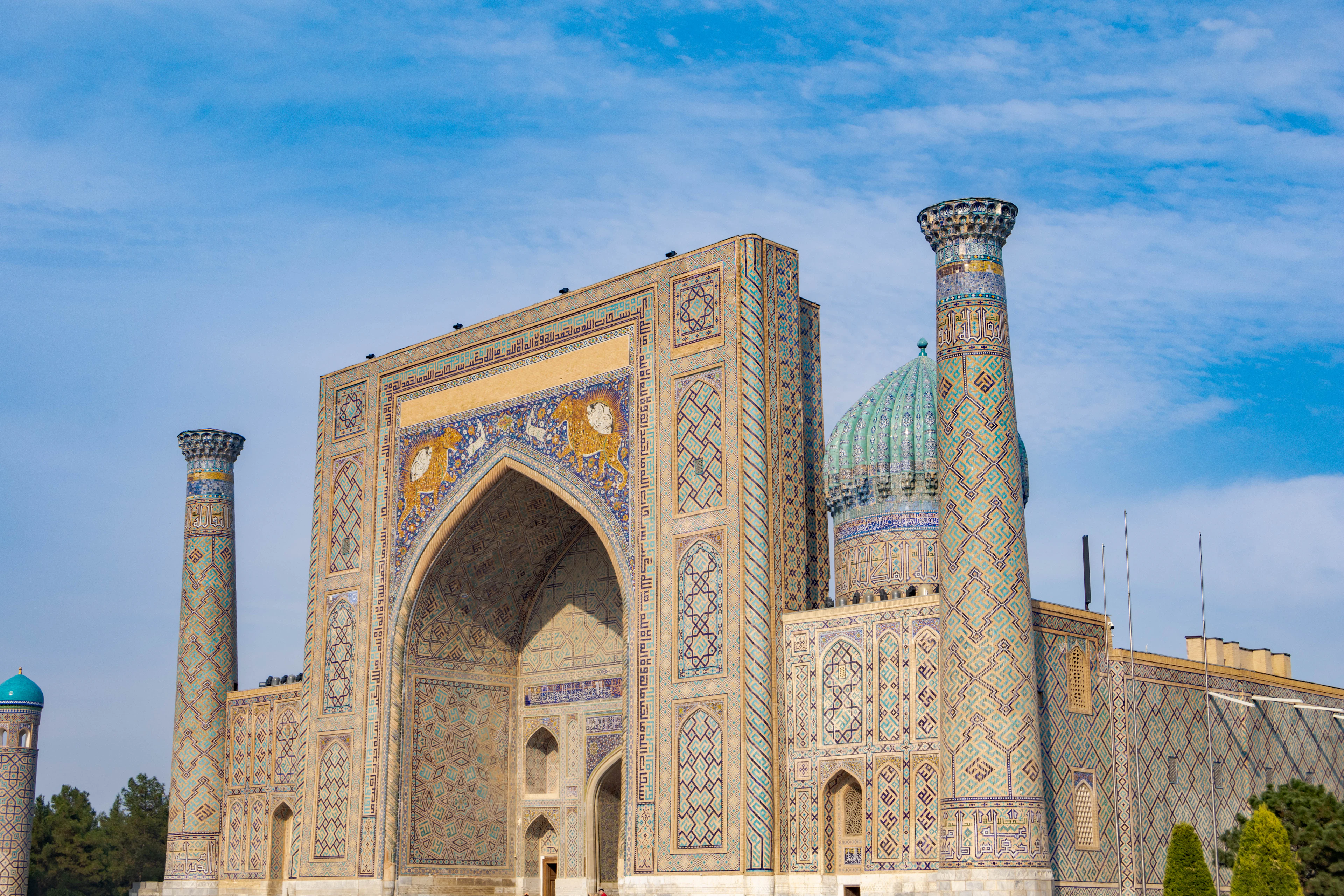 The Sher-Dor Madrasah at the Registan in Samarkand, its great mosaic portal flanked by ribbed minarets under a blue sky