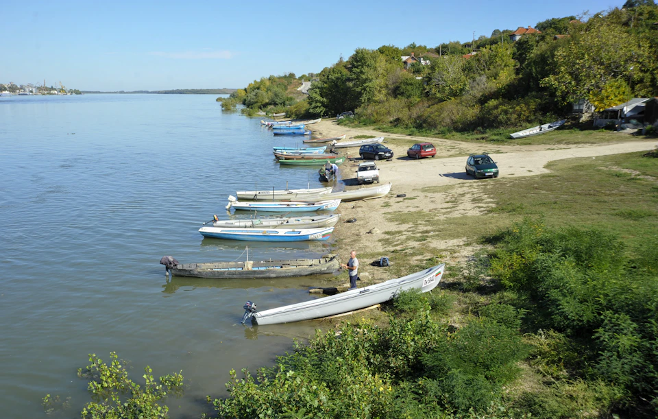 The image depicts a serene river scene with several boats docked along its banks, including white and blue boats, some moored to the shoreline while others are floating on the water's surface. The calm waters reflect the clear sky above, creating a tranquil atmosphere. In the foreground, there is a grassy area where people can be seen walking around or standing near their boats.

In addition to the boats and people, there are also cars parked nearby, suggesting that this location might be a popular spot for boating activities or recreational outings. The overall scene conveys a sense of relaxation and enjoyment as people spend time by the river with their boats.