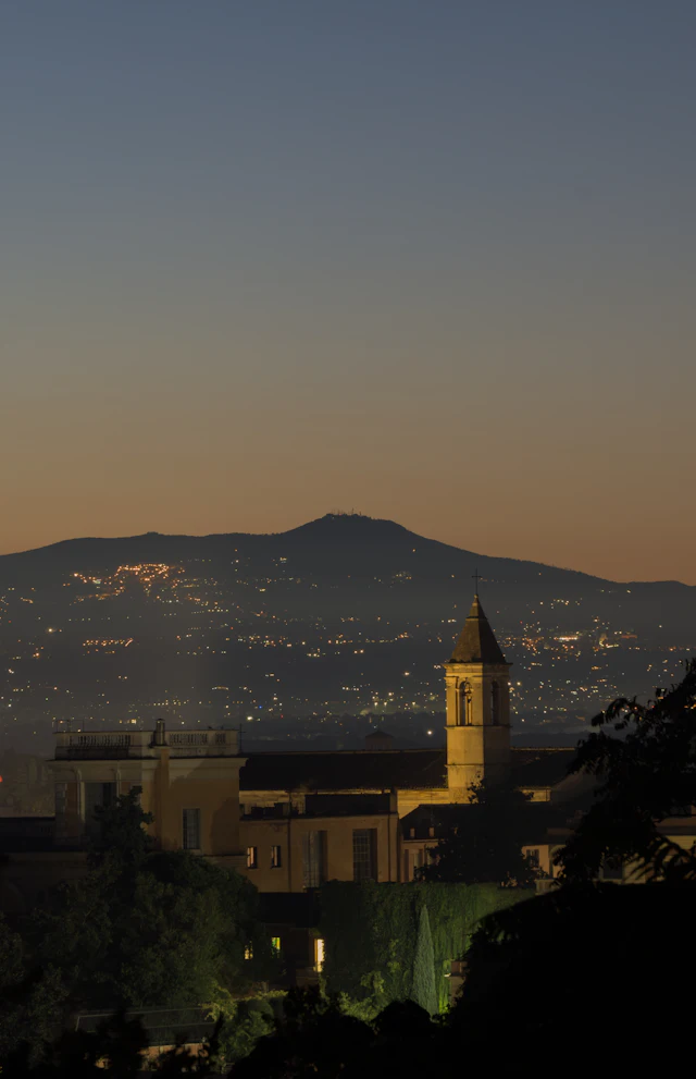 A dusk view over Rome's rooftops and hills from the Gianicolo, with a church bell tower silhouetted against the sky