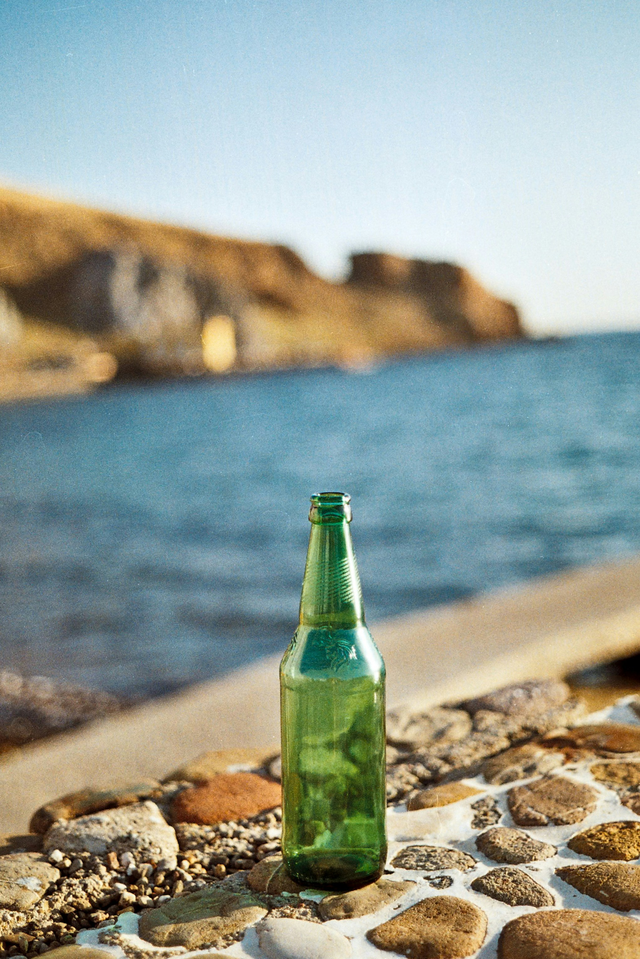 Green glass bottle sits on a pebbled shoreline with blue water and distant cliffs in the background.