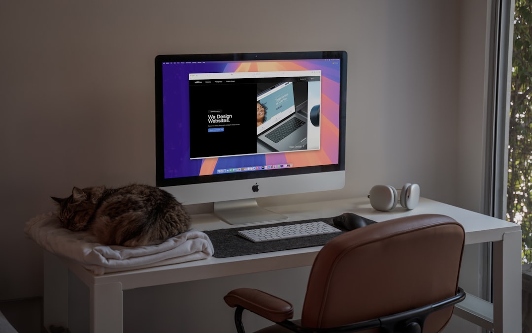 A cat laying on a desk next to a computer, A peaceful home office setup featuring an iMac showcasing a modern web design project, with a comfortable brown leather chair, wireless headphones, and a sleeping cat on a soft blanket. The natural light filtering through the window adds a calming atmosphere, making this an ideal space for creativity and productivity. Perfect inspiration for remote workers, designers, or anyone who loves minimalist workspaces with a touch of coziness.