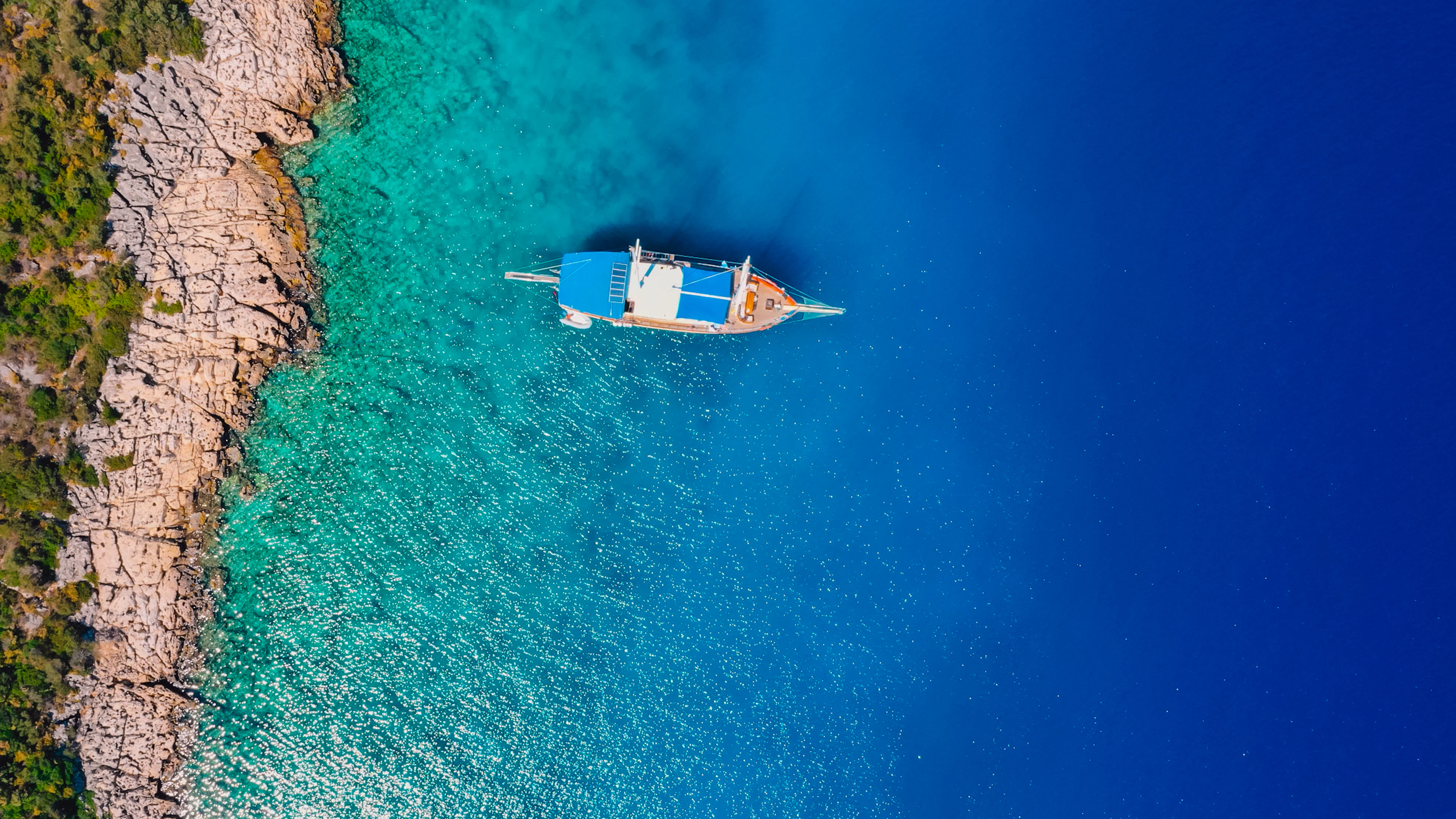 A wooden gulet in clear turquoise water off a rocky shore near Kaş, seen from above