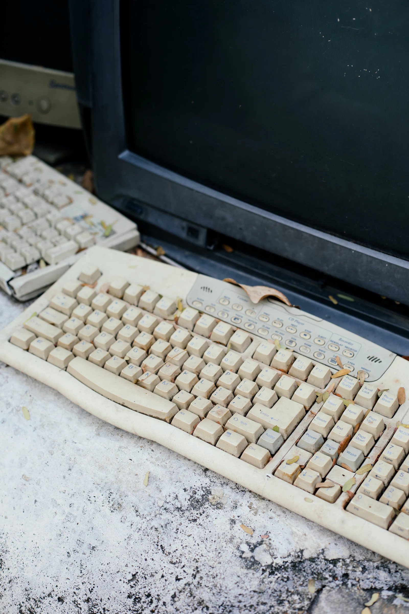 A yellowed beige keyboard and monitor left outdoors, dusty and leaf strewn.
