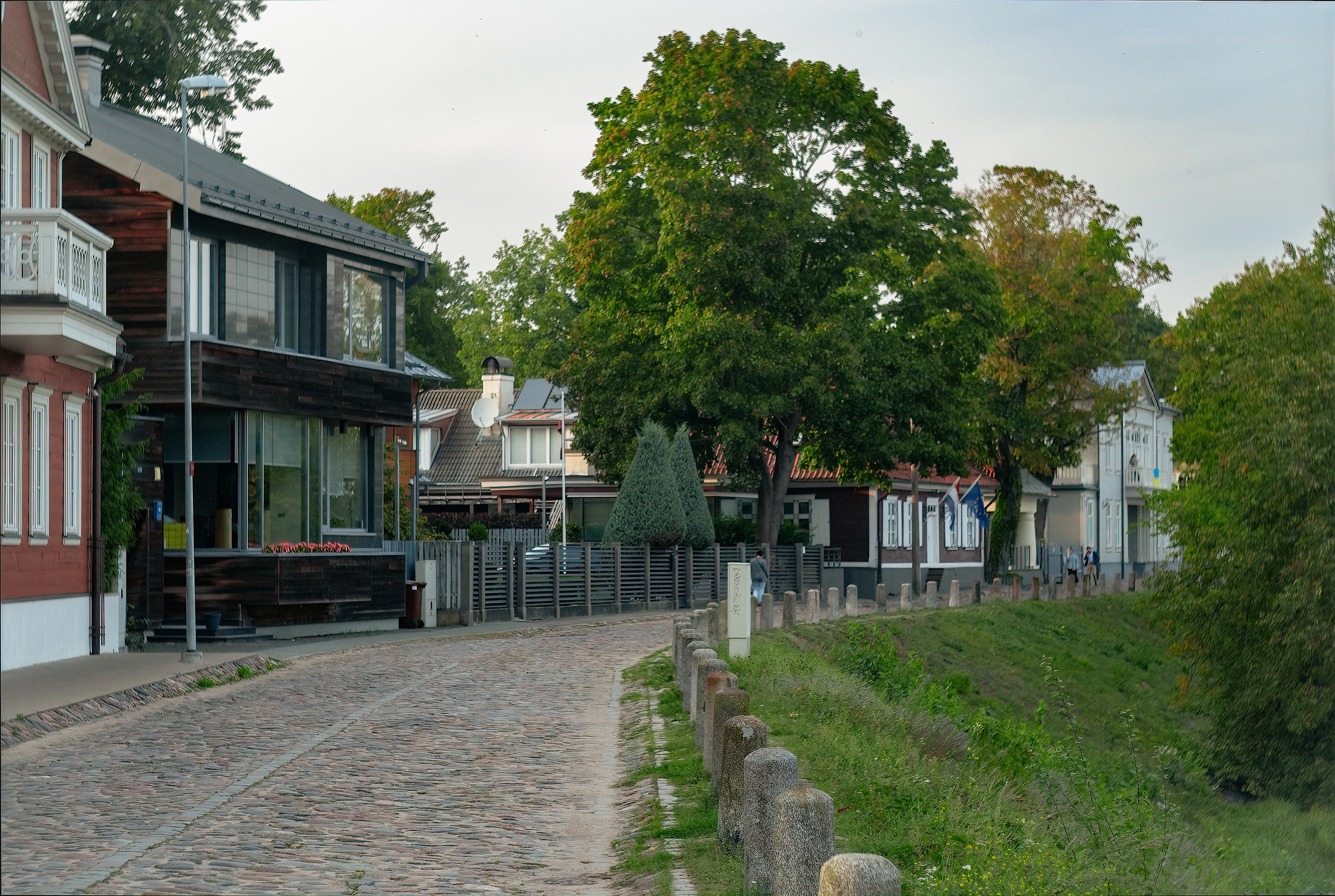 A cobbled street on Ķīpsala island in Riga lined with painted timber houses and a grassy embankment, its old stone bollards running away under soft grey evening light.