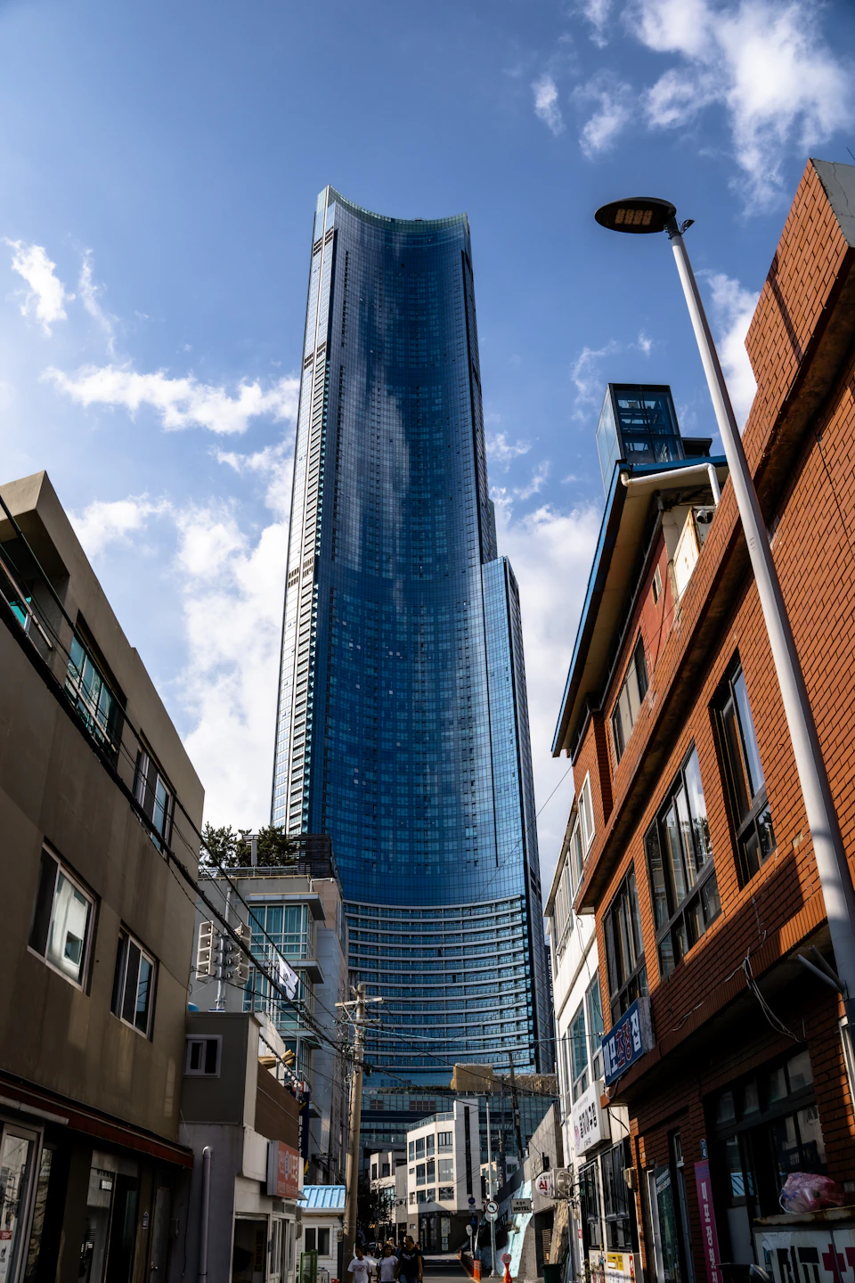 The image depicts a bustling city street with tall buildings lining both sides, including a prominent blue glass skyscraper on the right side and a brick building on the left. The sky above is clear blue with scattered clouds, creating a bright and sunny atmosphere. On the ground level, there are several people walking along the sidewalk, going about their day.

In addition to the buildings, there are also street lamps visible in the scene, one of which is located near the brick building on the left side of the image. The overall composition suggests an urban environment with a mix of architectural styles and daily life activities taking place.