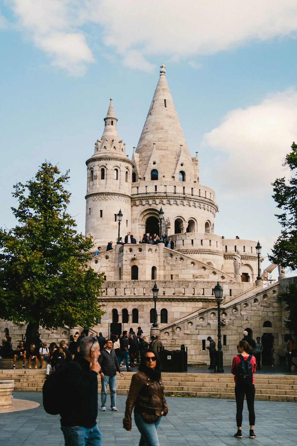 The image depicts a large stone castle with multiple towers and spires, standing majestically against a clear blue sky. The castle is situated on a hill, providing an impressive view from its vantage point. A group of people can be seen walking around the base of the castle, adding life to the scene. Some individuals are carrying backpacks, suggesting they may be tourists exploring the area or locals going about their daily routines.

The image also contains some text that reads "sa_1896".
