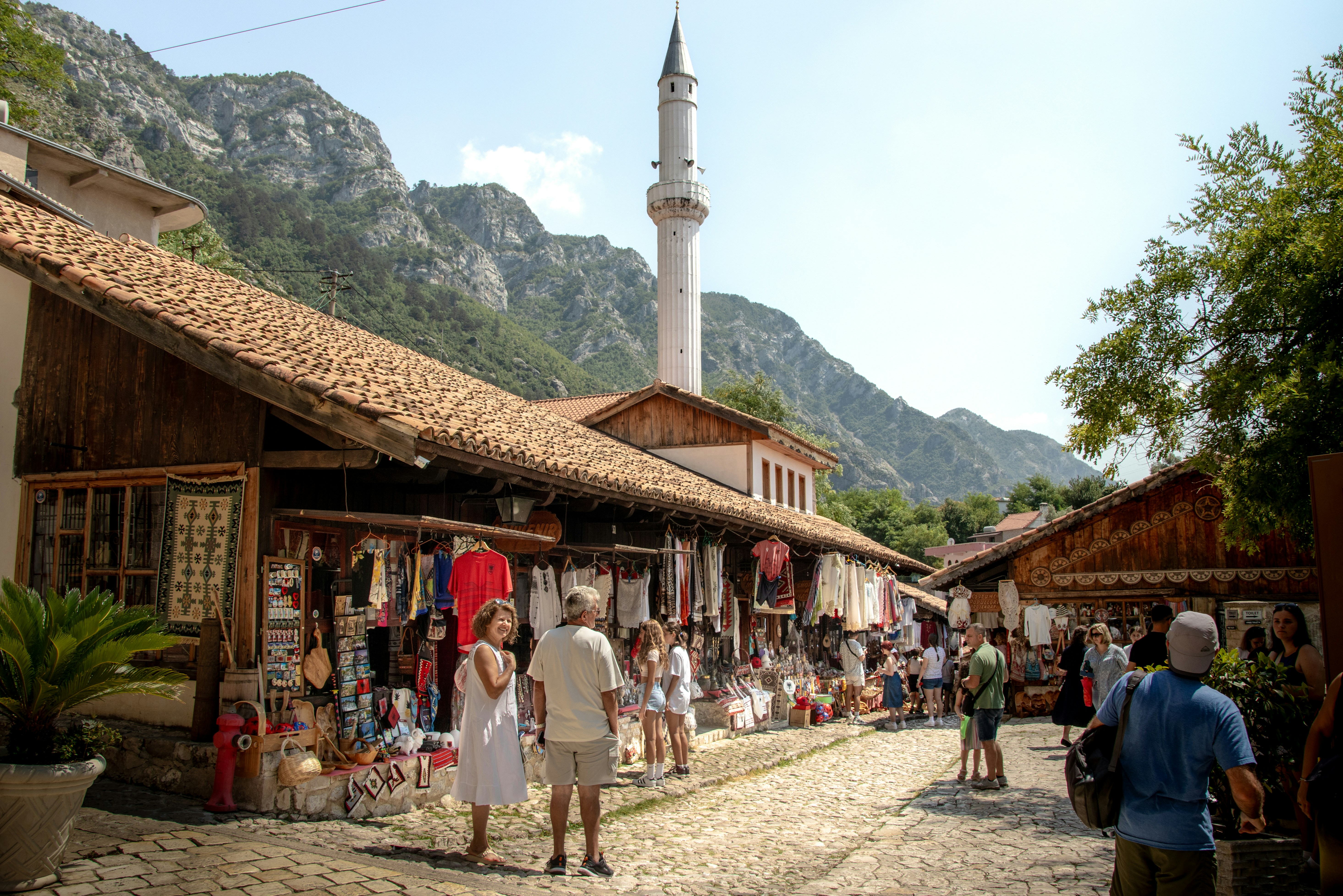 A group of people walking down a street next to a tall building, Albanian market in Krujë.