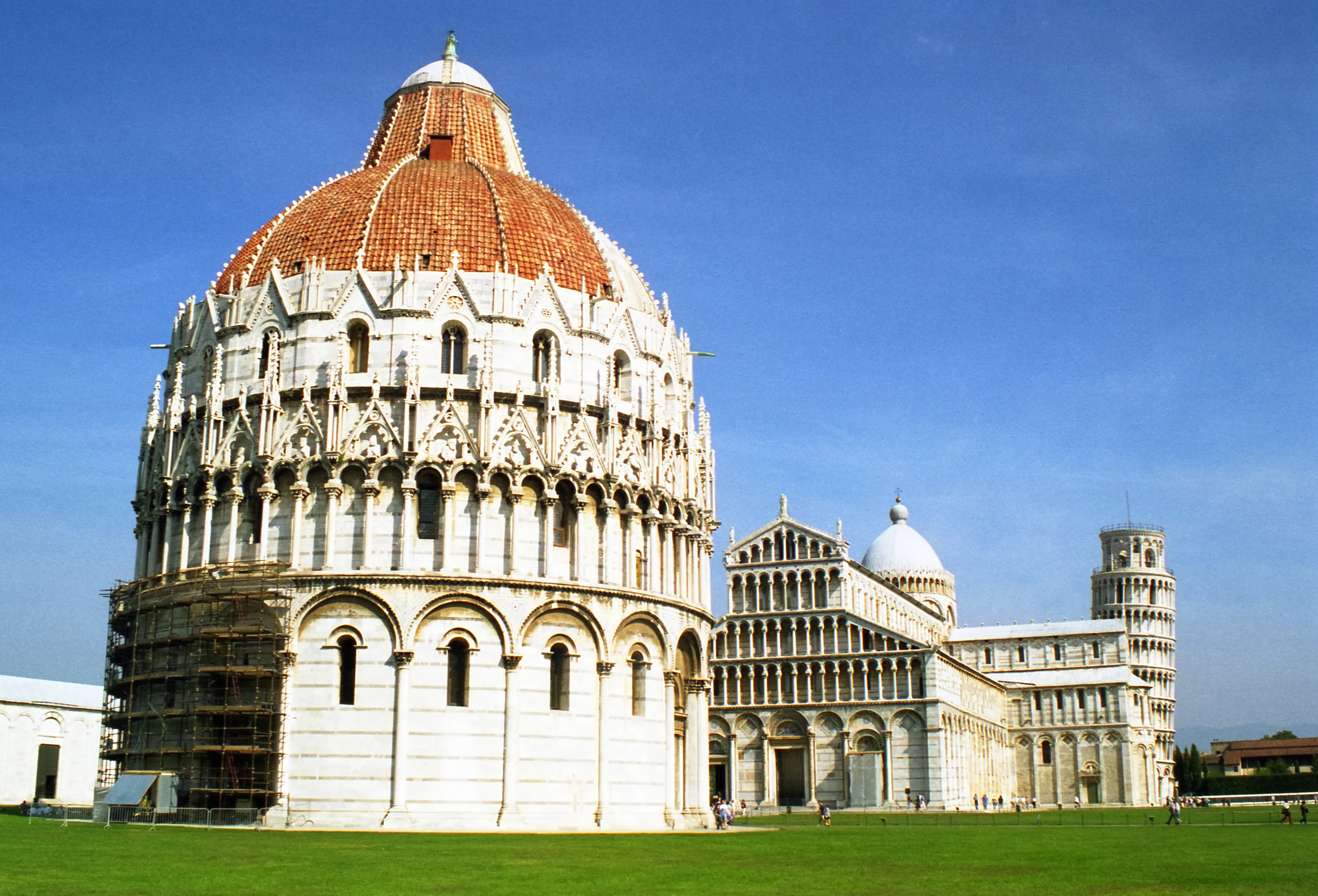 The Baptistery, Cathedral and Leaning Tower of Pisa together on the green lawn of the Piazza dei Miracoli under a blue sky.