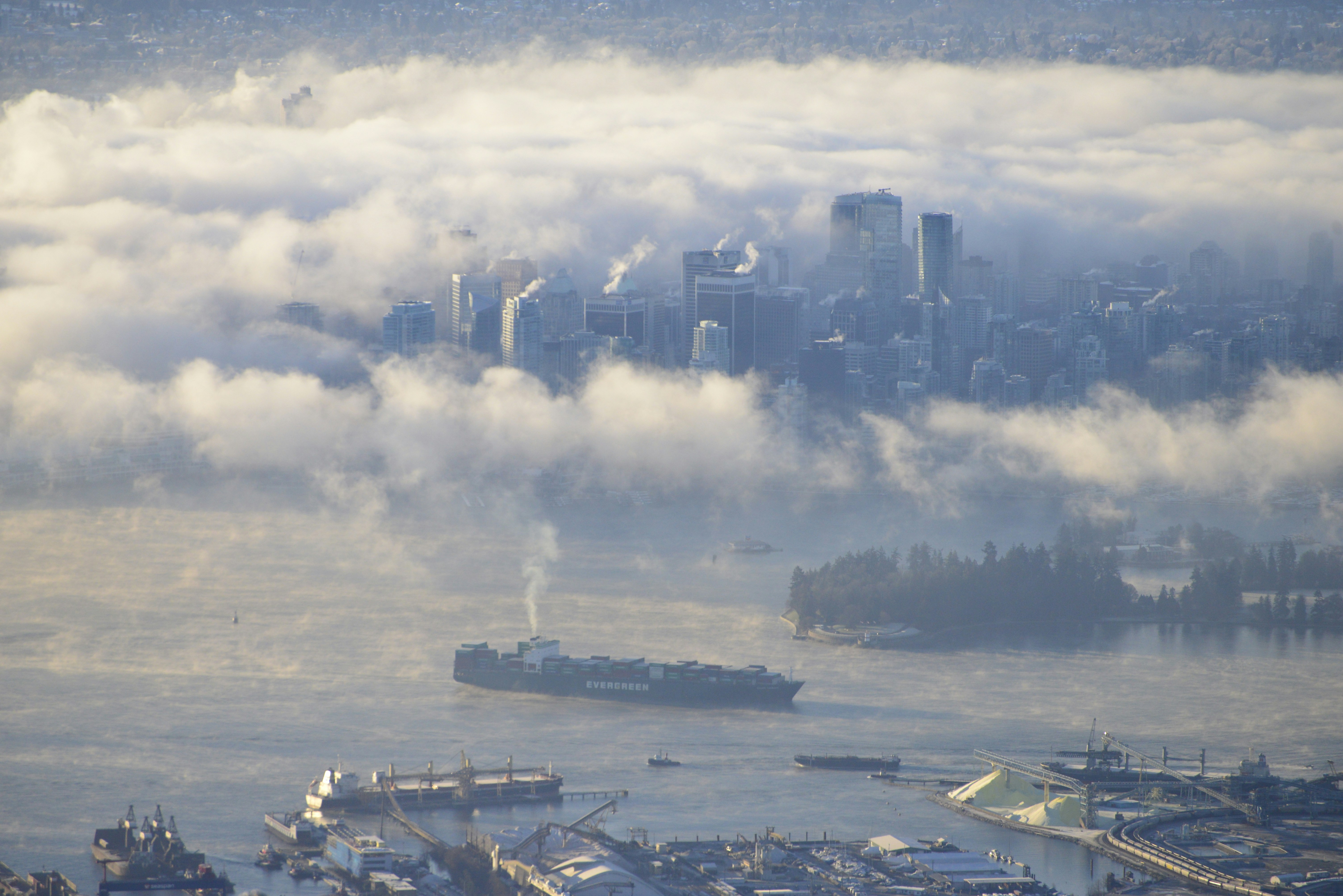 Downtown Vancouver's skyline emerges above low cloud and fog, with a cargo ship and mountains in the harbour below