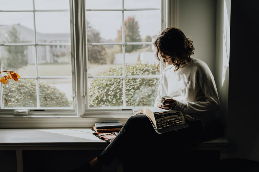 An adult in a cream sweater sits in a sunlit window nook with a warm drink, head bowed in a quiet reflective moment