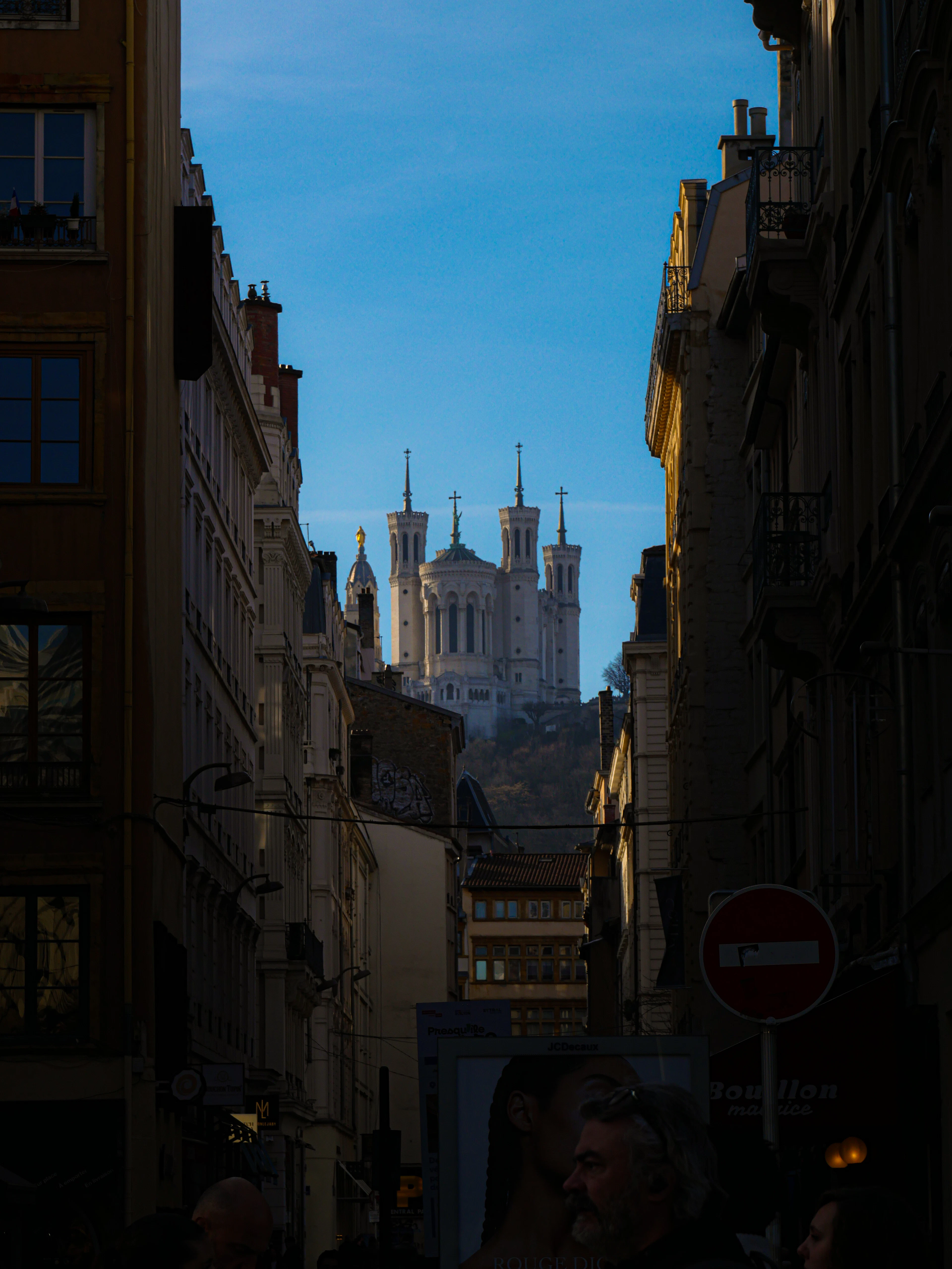 The four white towers of the Basilica of Notre-Dame de Fourviere rise above the hillside at the end of a narrow, deeply shadowed Lyon street under a clear blue winter sky.