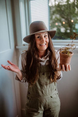 A woman wearing overalls and a hat holding a potted plant