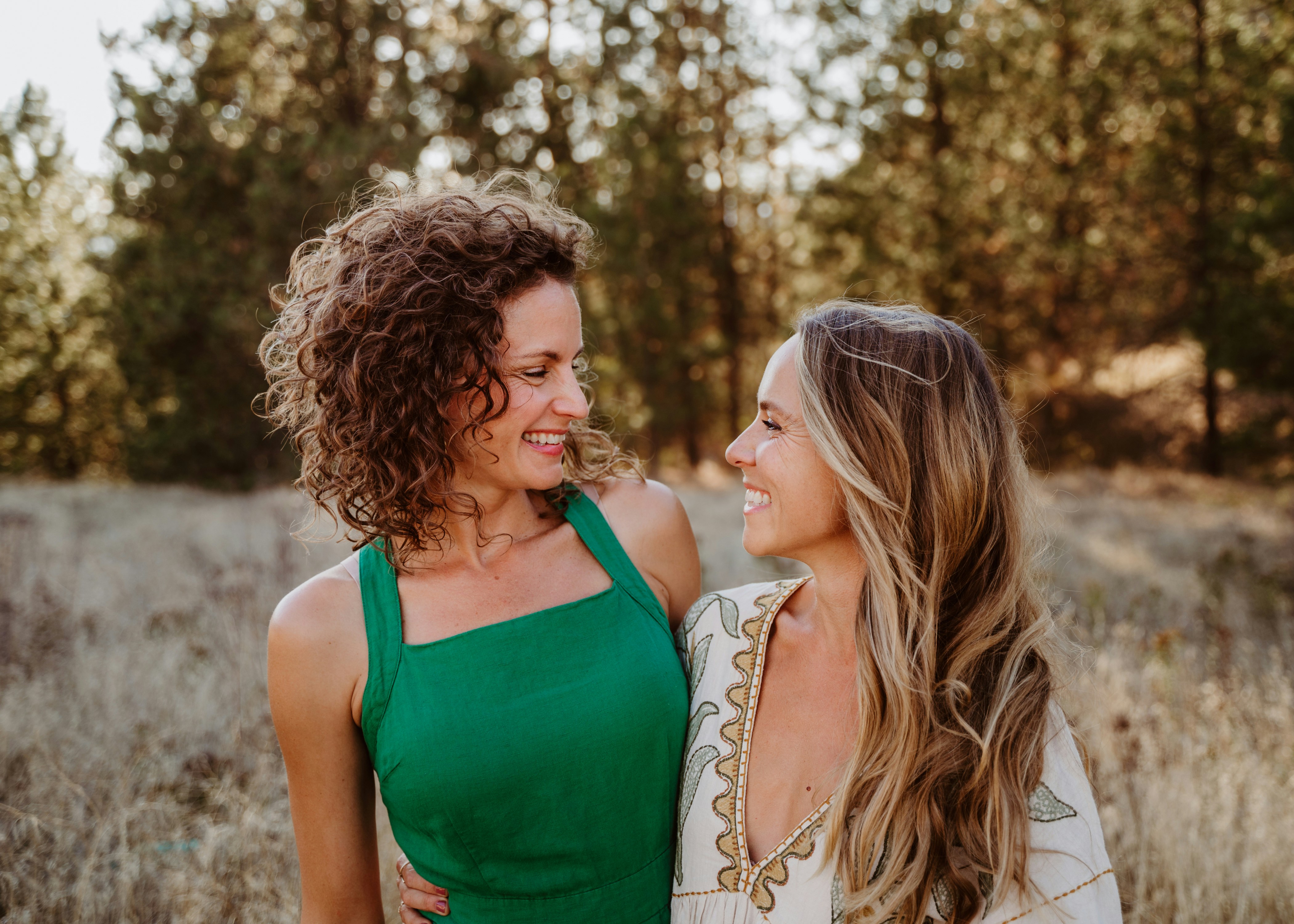 Two women embracing and smiling in a grassy meadow surrounded by evergreen trees