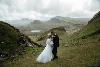 A bride and groom standing on a grassy hill