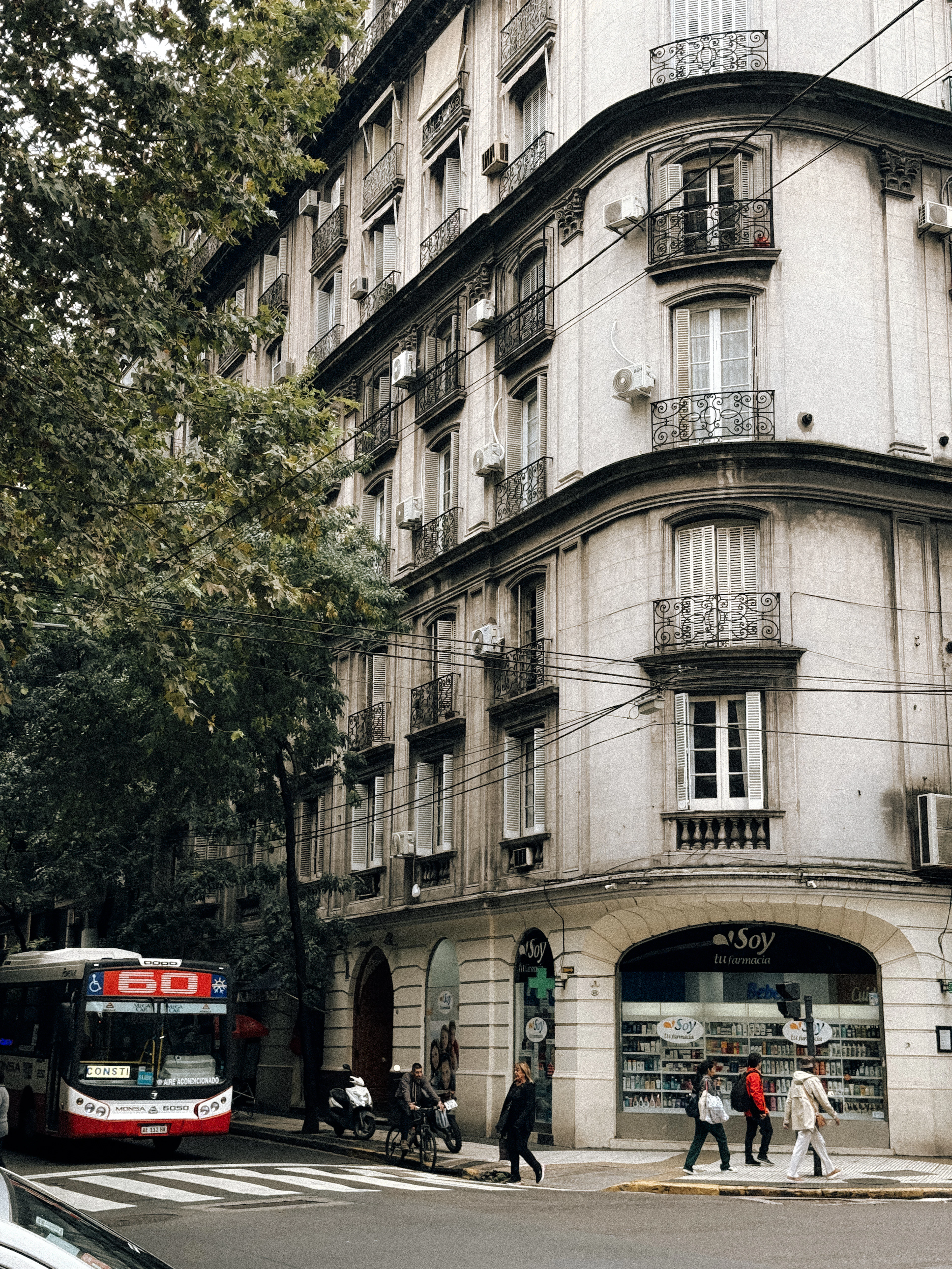A red-and-white number 60 colectivo pulling past a curved Belle Epoque corner block in Buenos Aires, tiers of wrought-iron balconies above a corner pharmacy while pedestrians cross in front.