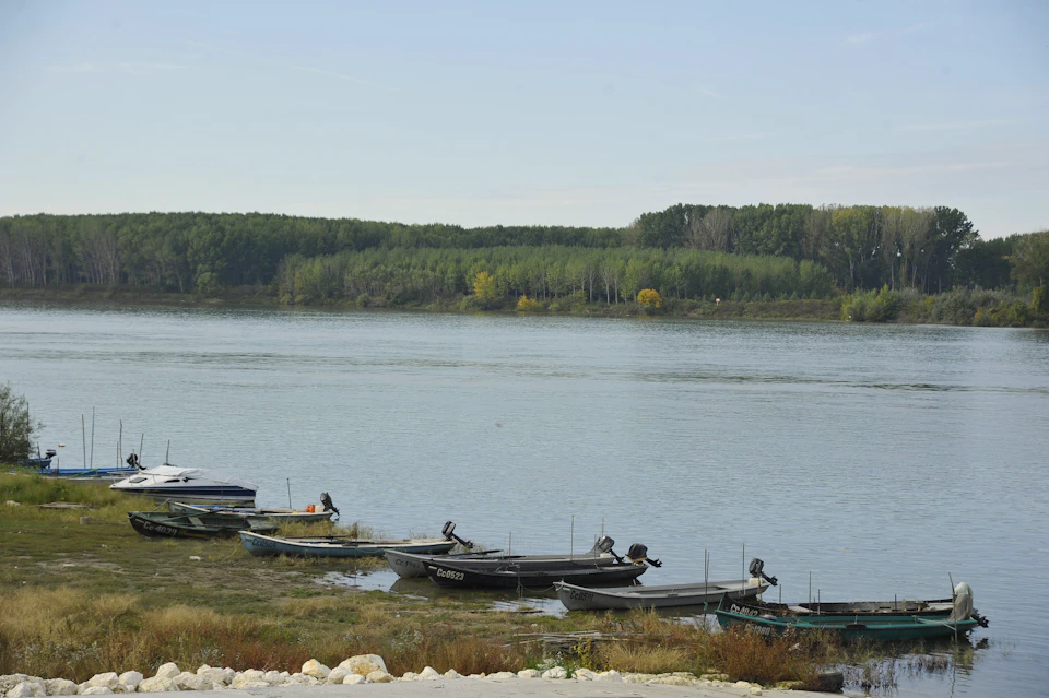 The image depicts a serene lake scene with several boats docked along its shoreline. The calm water reflects the clear blue sky above, creating a tranquil atmosphere. On the left side of the image, there are six boats moored to the shore, including two motorboats and four smaller boats. In the background, on the right side of the image, there is a line of trees standing tall against the horizon.

The perspective of the photo is from above, providing an expansive view of the lake and its surroundings. The sky forms the backdrop for this picturesque scene, enhancing the overall beauty of the landscape.