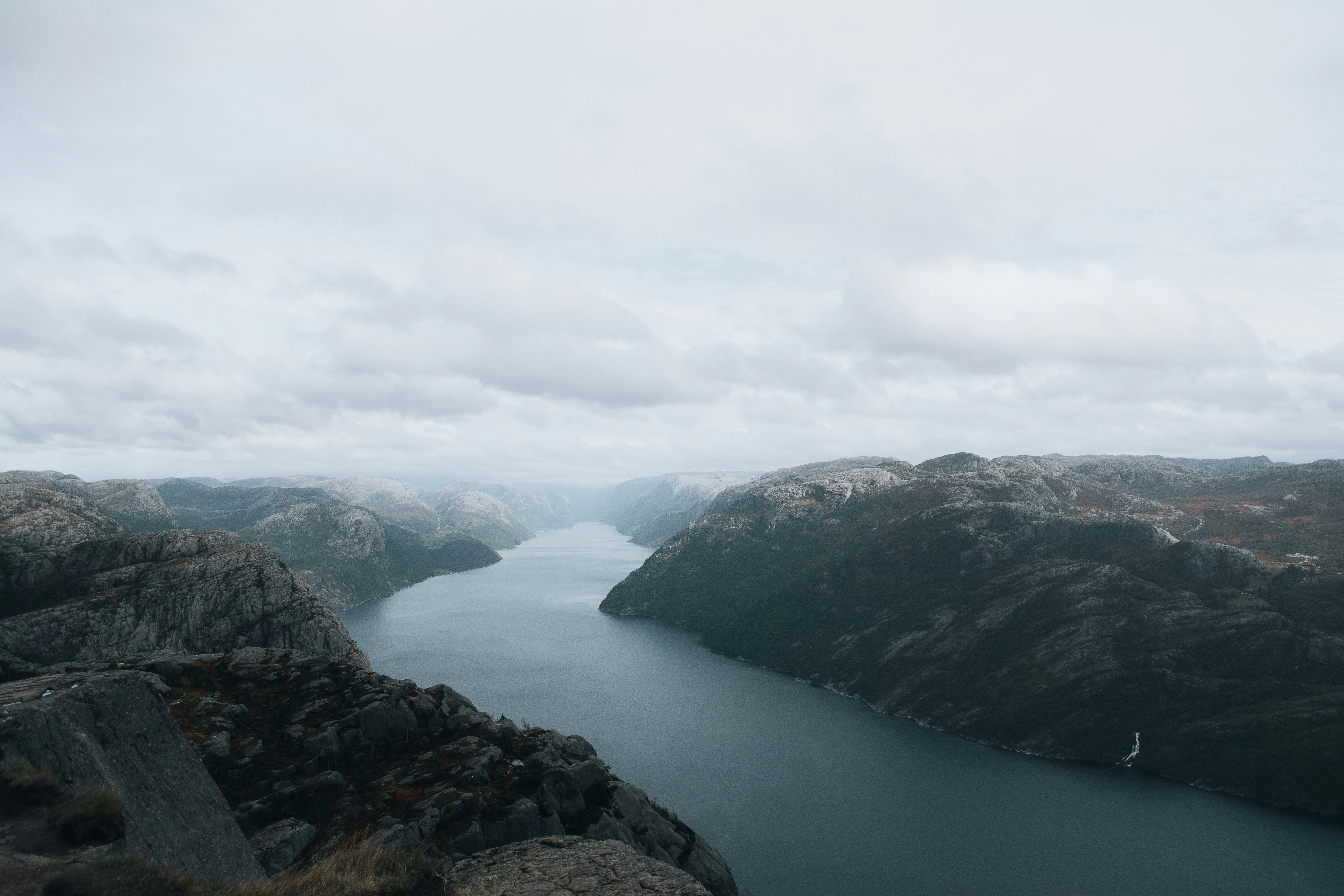 A panoramic view of a fjord with steep, rocky cliffs extending down into the calm water below. The overcast sky casts a soft light on the rugged terrain, emphasizing the dramatic landscape. The distant mountains blend into the horizon, while the waterway meanders through the valley, creating a serene and remote atmosphere.