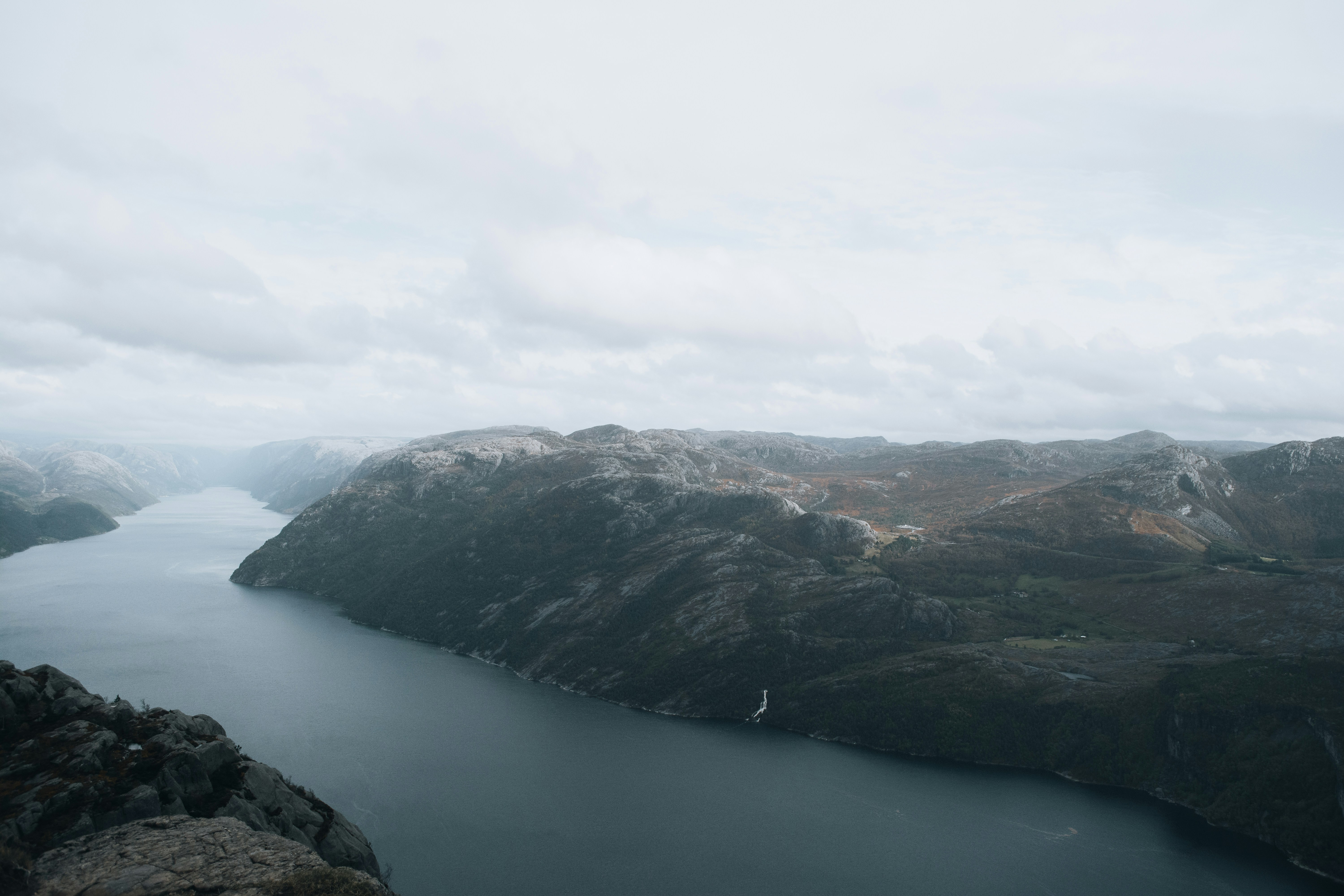 A panoramic view of a fjord with steep, rocky cliffs extending down into the calm water below. The overcast sky casts a soft light on the rugged terrain, emphasizing the dramatic landscape. The distant mountains blend into the horizon, while the waterway meanders through the valley, creating a serene and remote atmosphere.