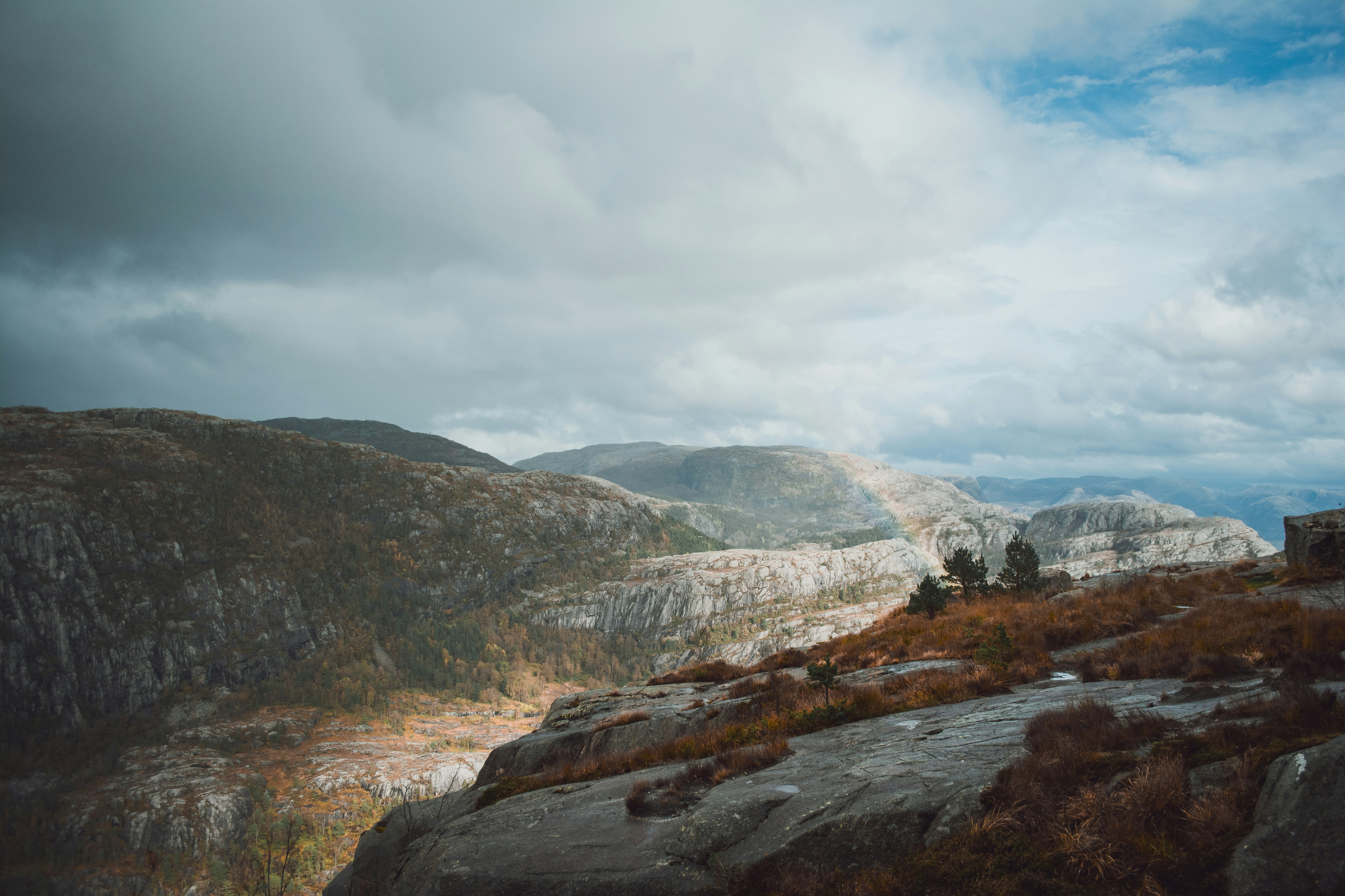 A wide landscape shot of rugged, rocky highlands under a moody, overcast sky. The rocky terrain features cliffs and outcrops, with sparse vegetation on the slopes. Patches of autumn colors can be seen in the distance, adding a touch of warmth to the otherwise dramatic and wild scenery. The cloudy sky casts soft light over the landscape, enhancing the sense of solitude and remoteness.