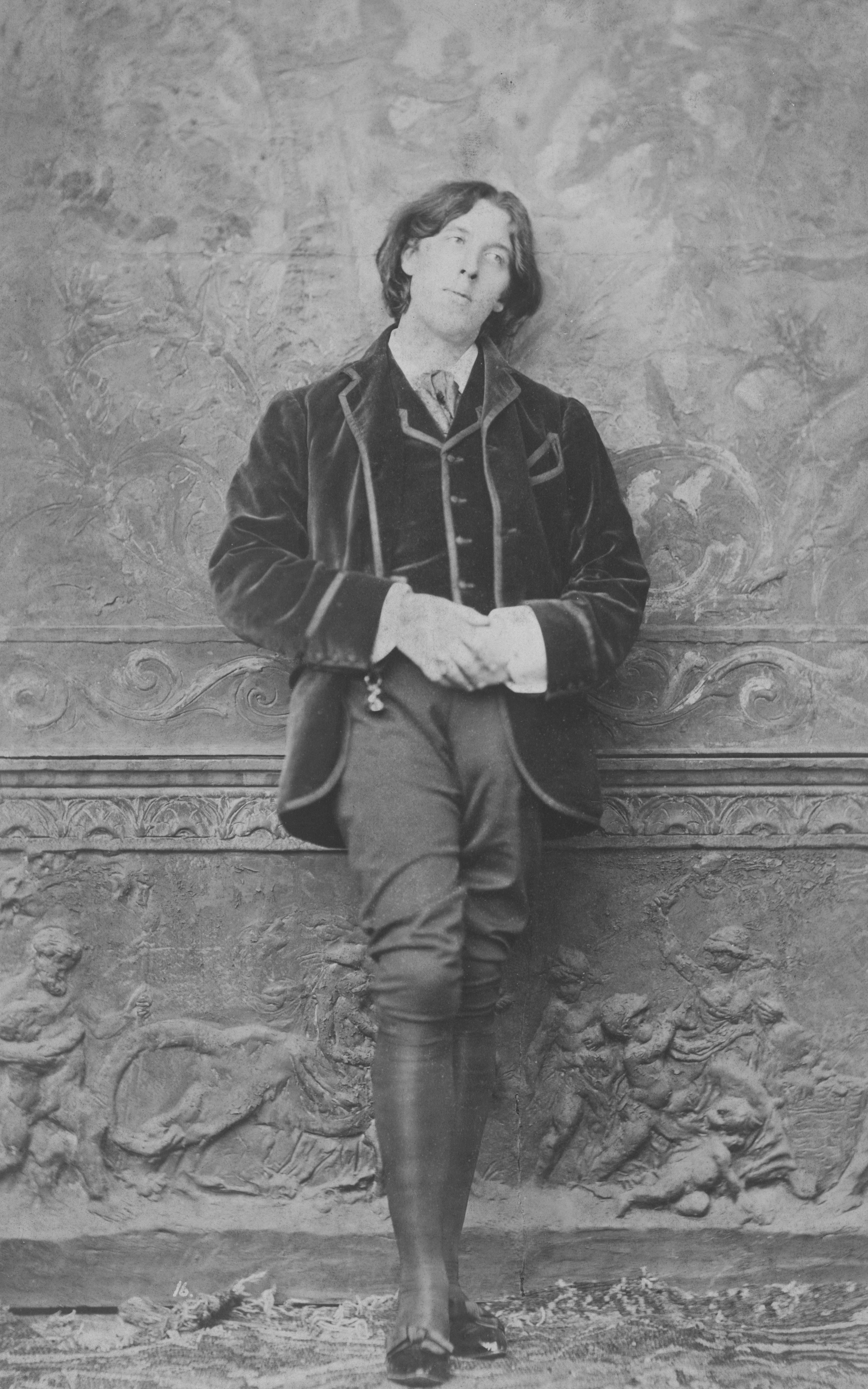A man in formal attire leans against a decorative wall.