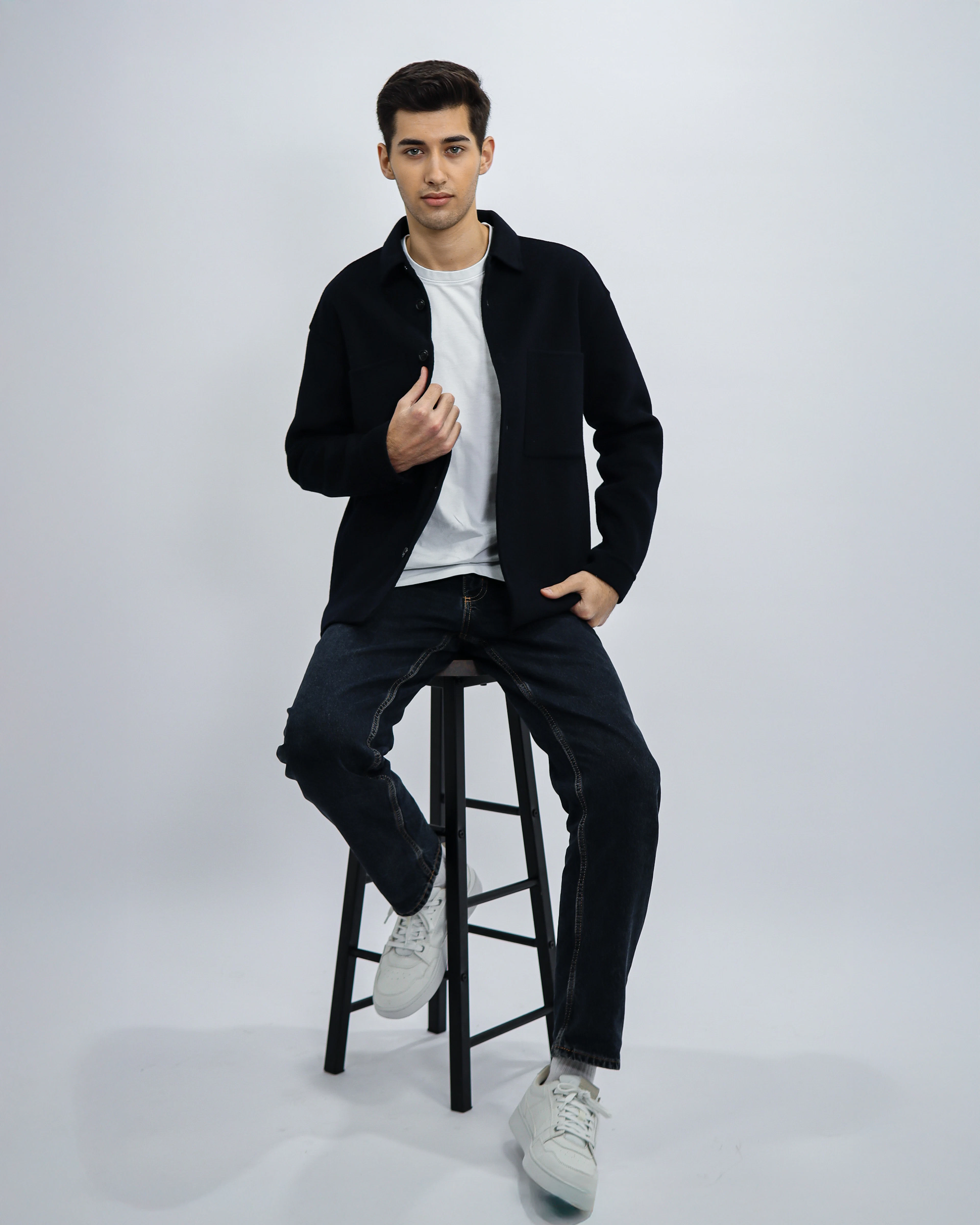Man in a black overshirt and white tee seated on a studio stool