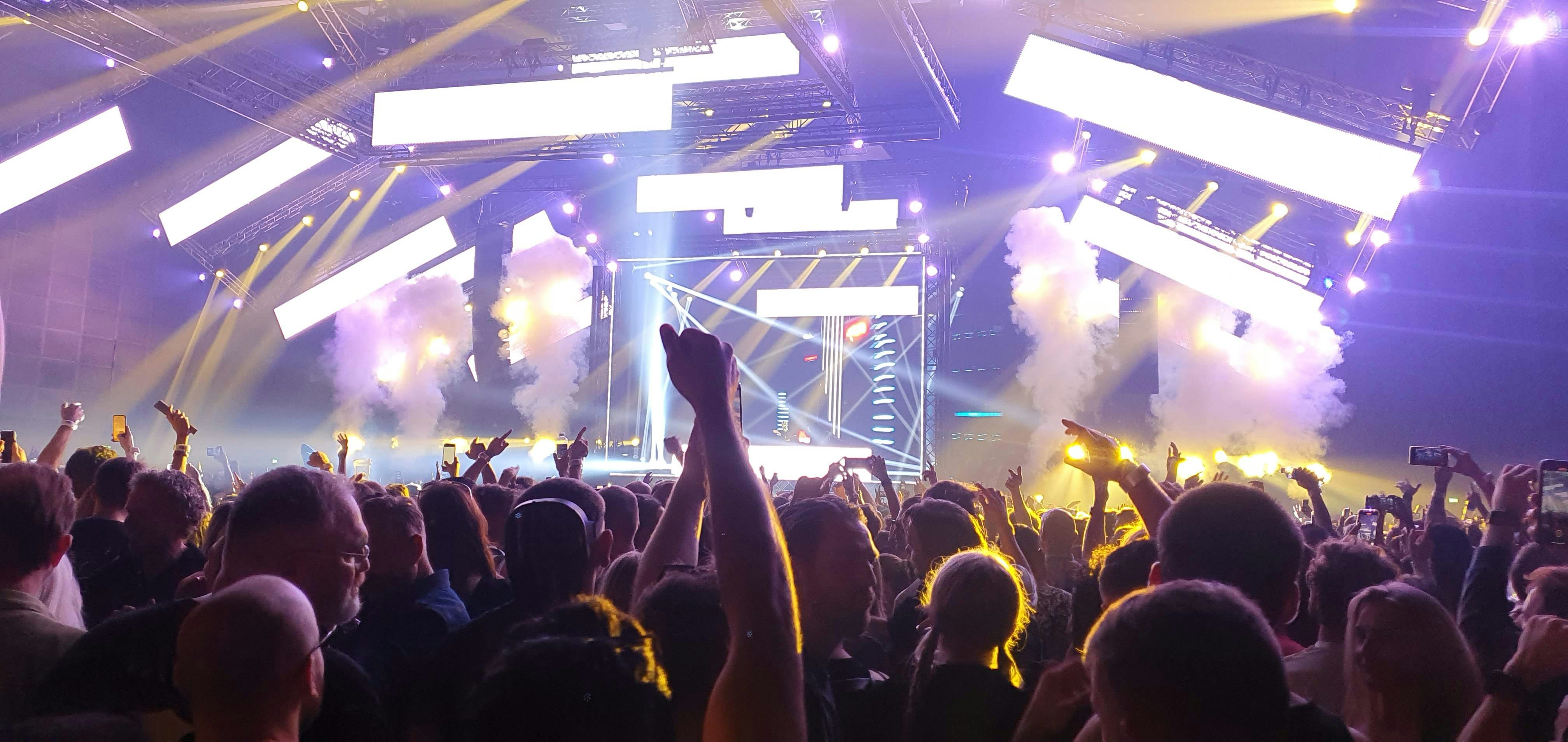 Live concert performance — packed venue with crowd, purple stage lighting, smoke effects, electric atmosphere