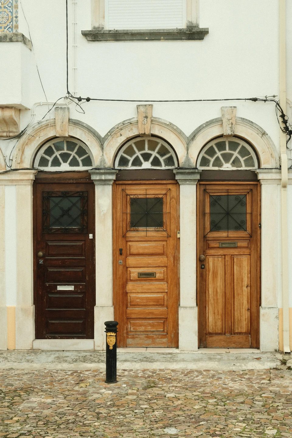 The image shows a row of three wooden doors, each with its own unique color and style, set against a white building facade. The first door is painted black and features a window above it. The middle door has a brown hue and also includes a window. The third door is the largest in size and is painted white, with a small window on the right side of the door.

In front of each door, there's a black trash can. The building itself appears to be made of stone or concrete blocks, giving it an old-world charm. A street lamp stands nearby, adding to the urban feel of the scene.