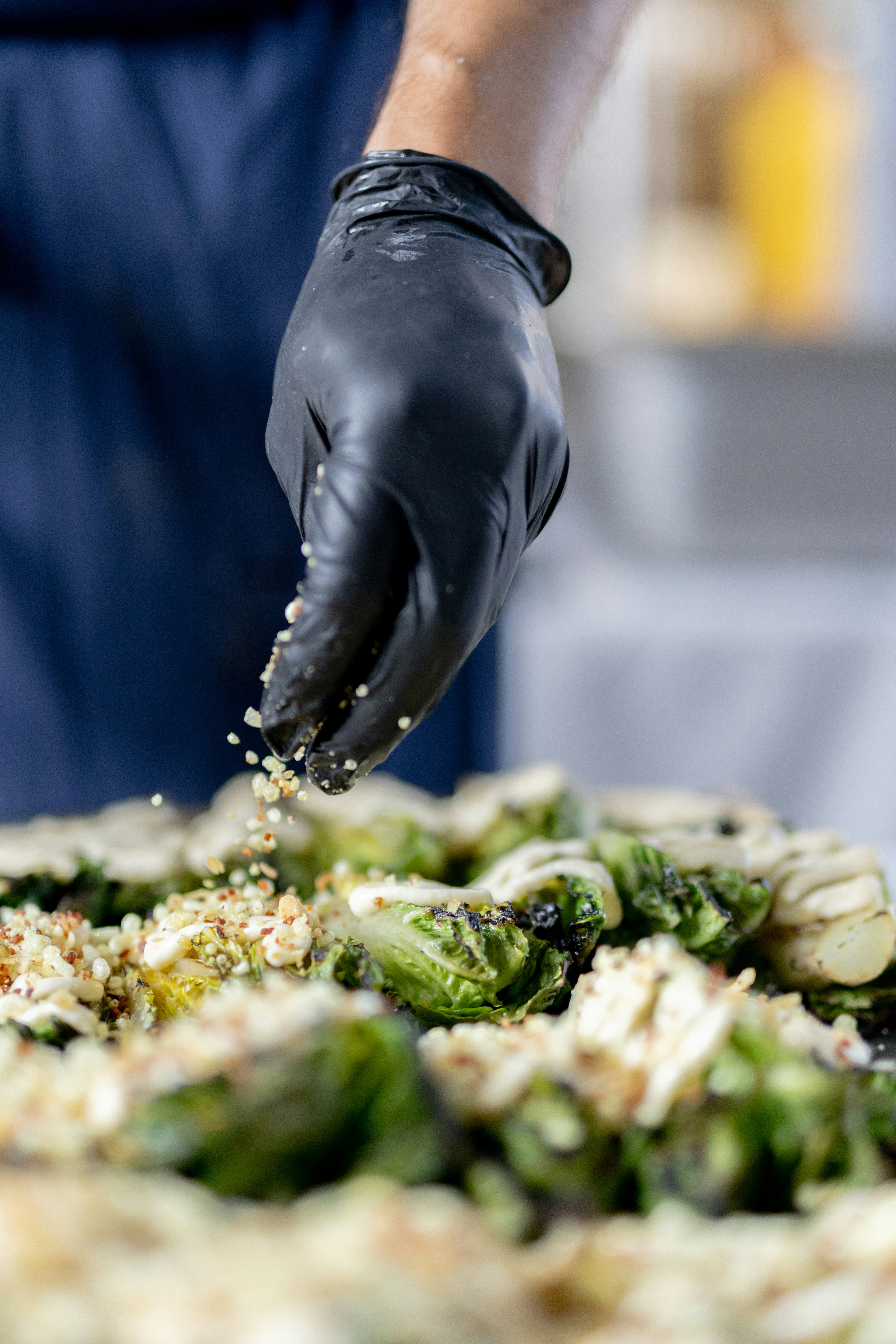 A person in a blue apron and black gloves is putting seasoning on a dish