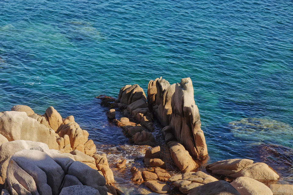 The image depicts a rocky coastline with a large, jagged rock formation on the left side and a smaller, smoother rock formation on the right side. The rocks are brown and gray in color, contrasting with the blue-green water that surrounds them. The sky above is clear and blue, suggesting a bright and sunny day.