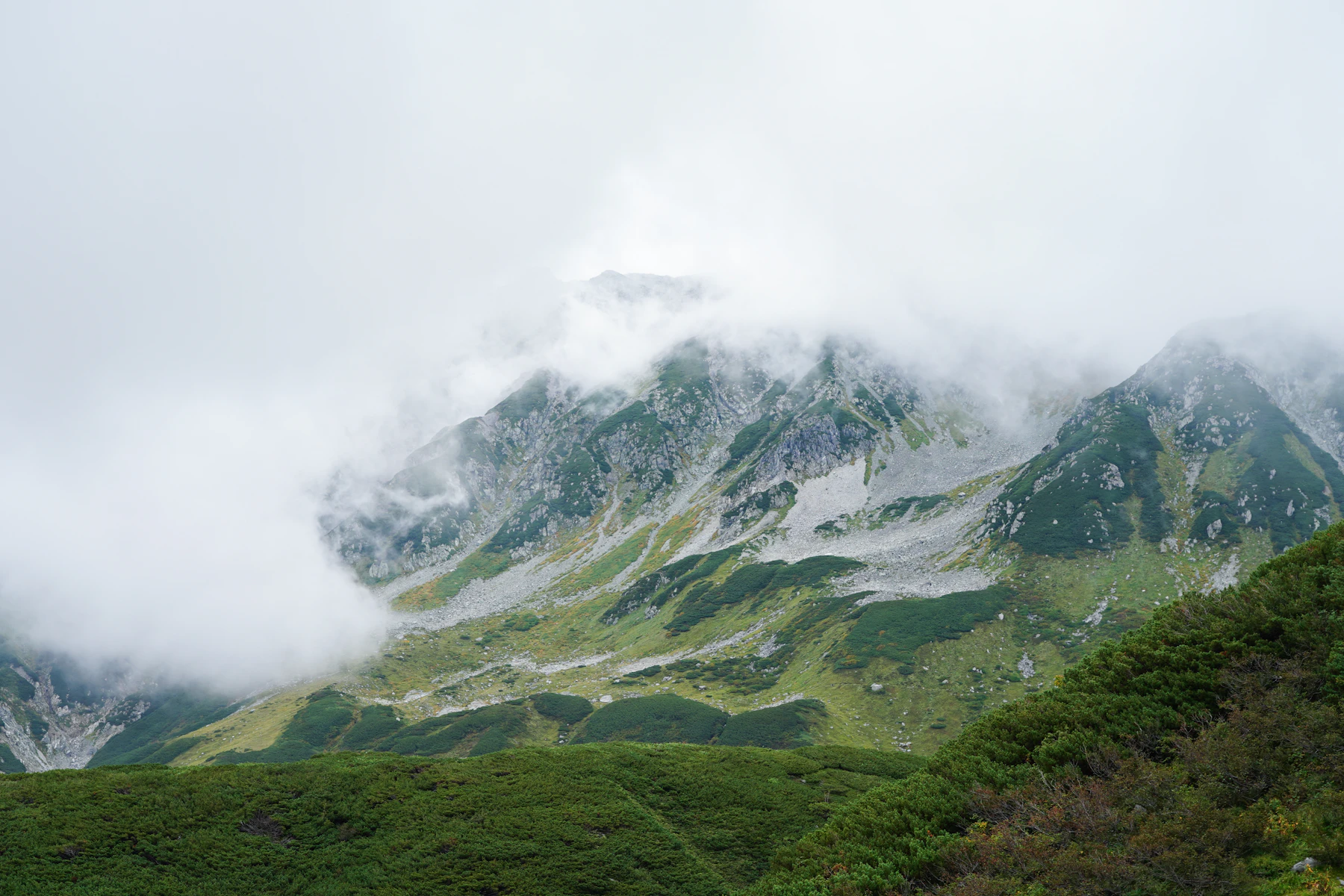 Cloud pouring over a green alpine ridge with snow on the tops