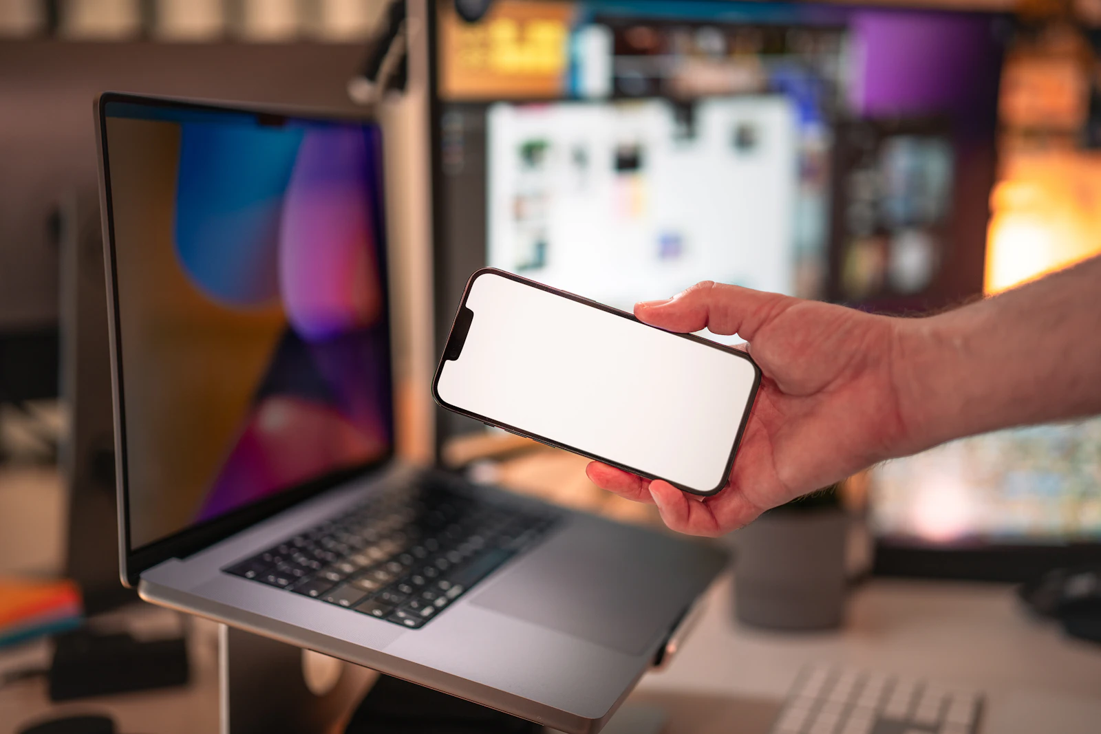 A hand holding a smartphone in front of an open laptop on a desk