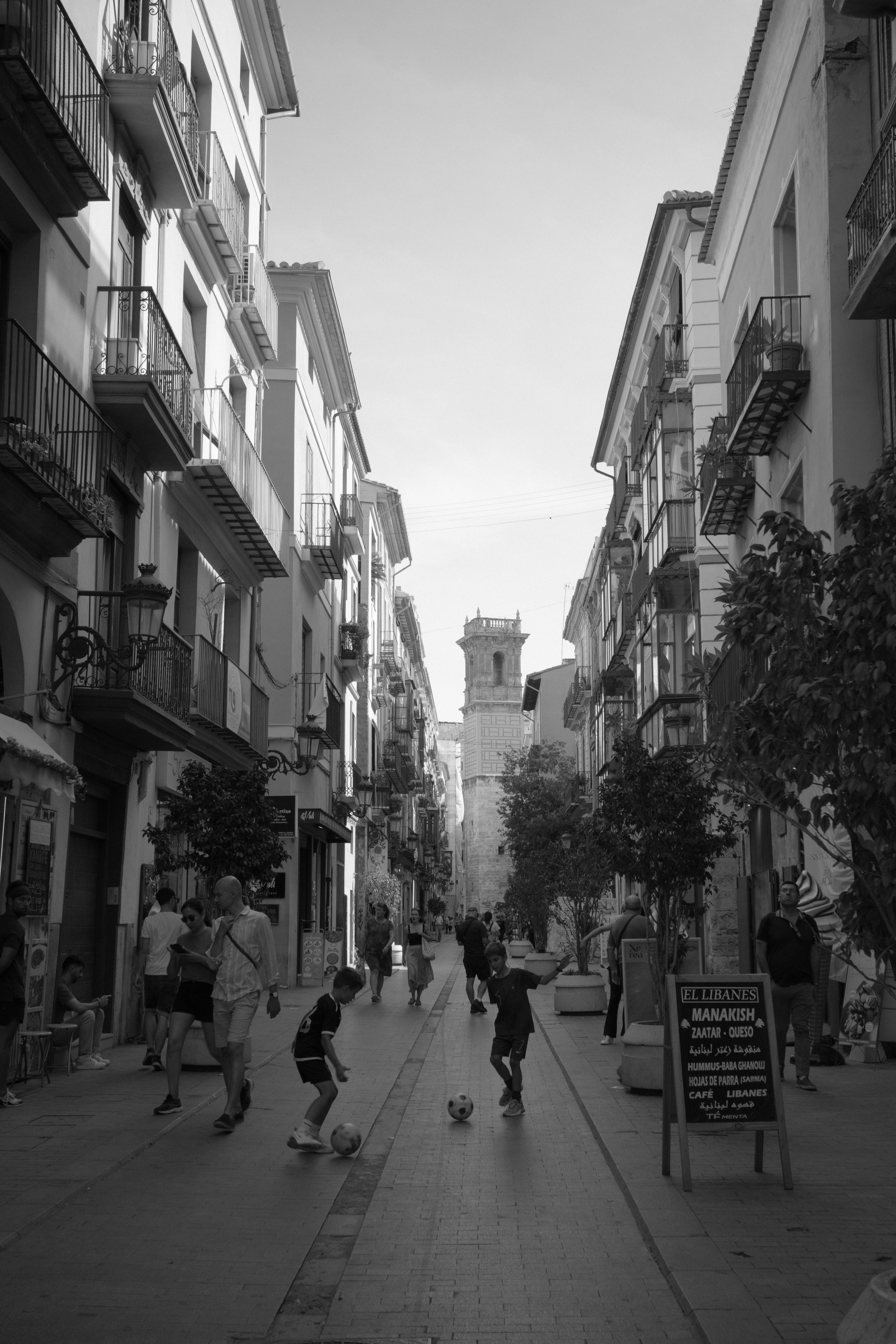 Kids play football down a narrow street of balconied buildings in Valencia's old town, a bell tower ahead.