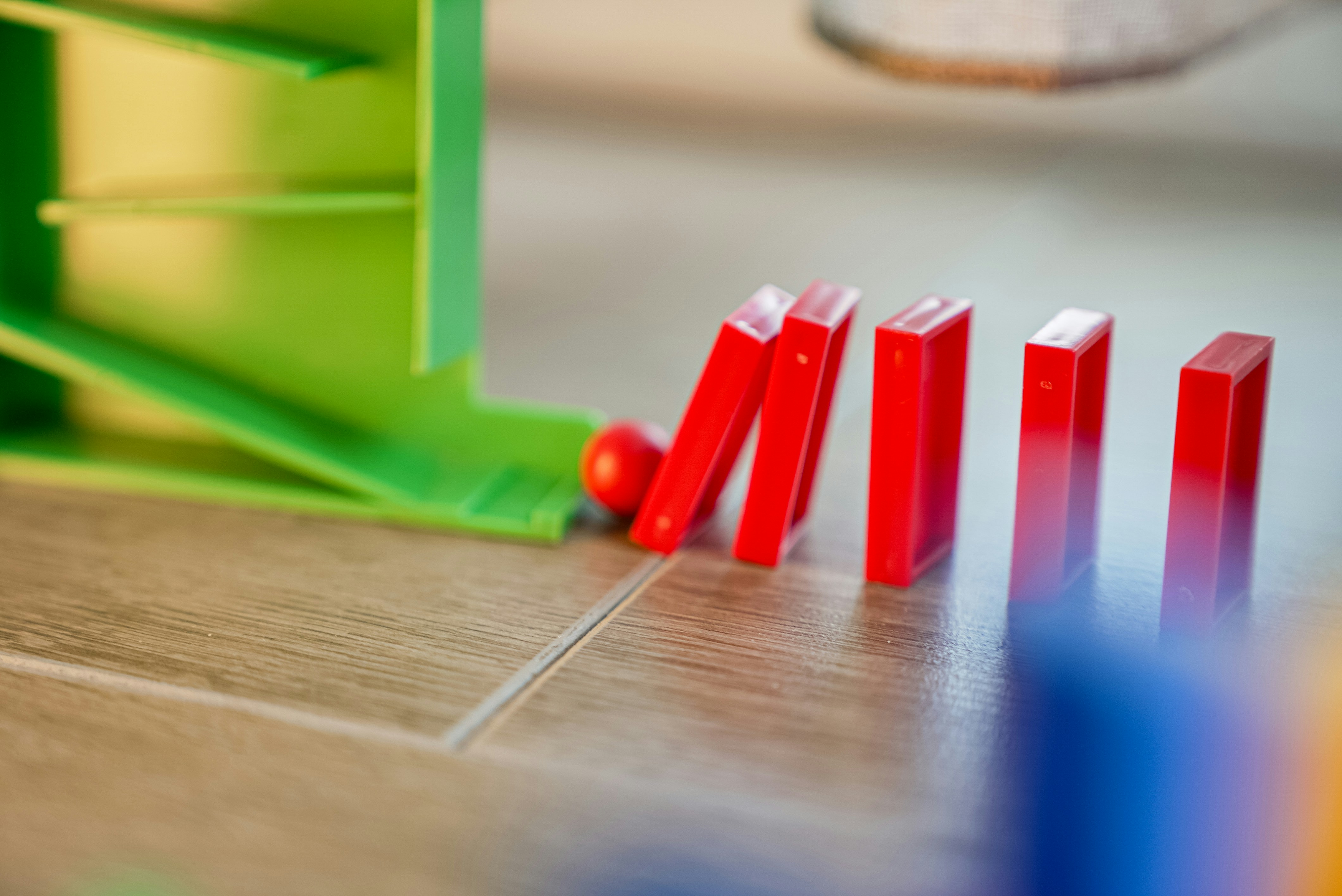 Red domino pieces falling in a sequence on a wooden surface, with a green toy structure in the background.