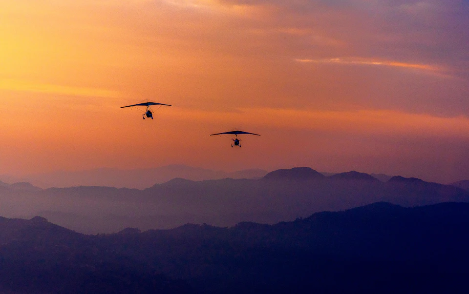 The image depicts two black and white paragliders soaring high above a mountainous landscape during sunset. The sky is painted with hues of orange, creating a stunning backdrop for the paragliders' flight path. One paraglider is positioned slightly ahead of the other, both appearing to be in mid-flight against this picturesque scene.

The mountains in the background are covered in trees and shrubs, adding depth and texture to the landscape. The sun's descent behind the mountains casts long shadows across the terrain, enhancing the overall beauty of the image.