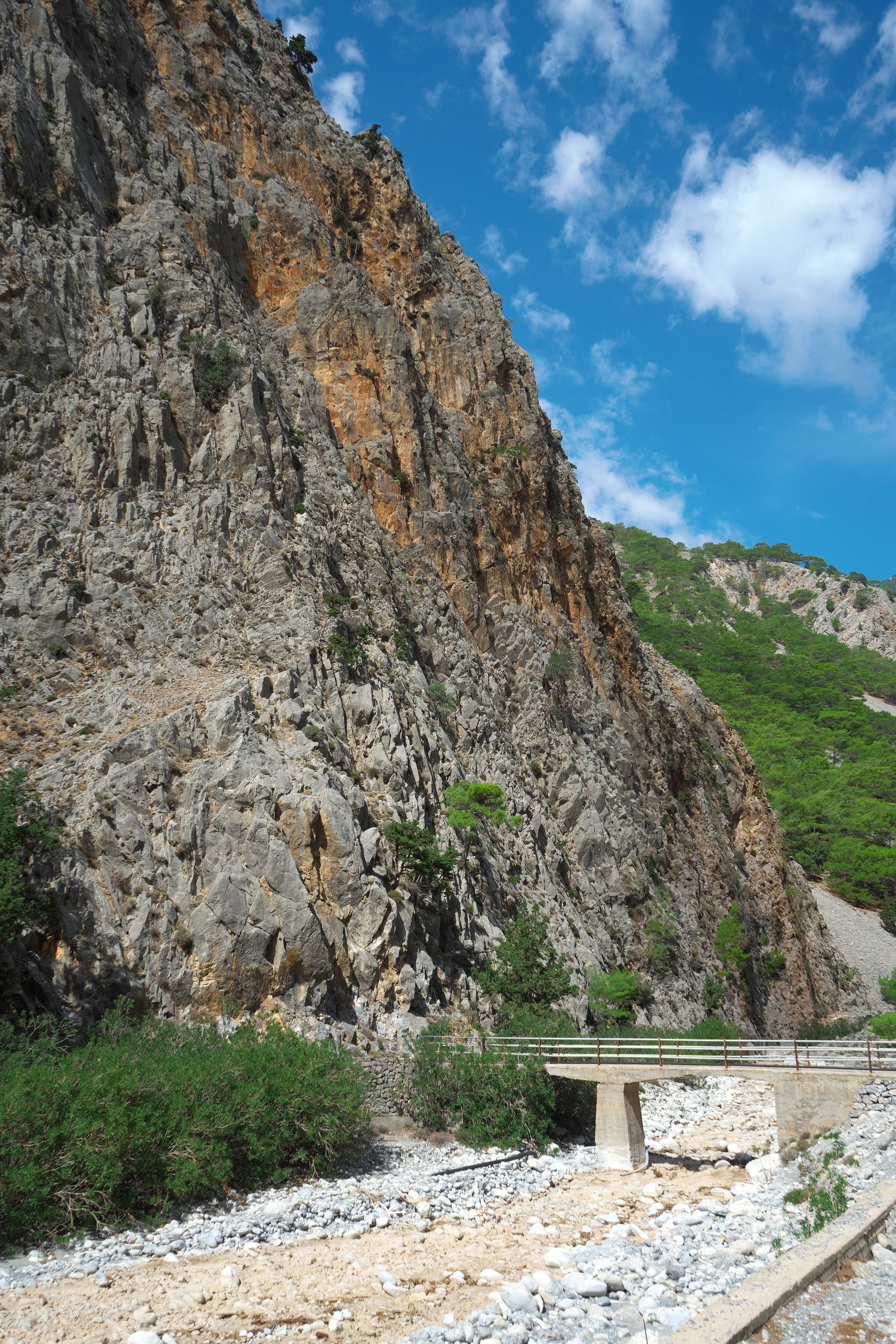 A rocky, sunlit gorge wall towers over a footbridge and dry streambed in the Cretan mountains.