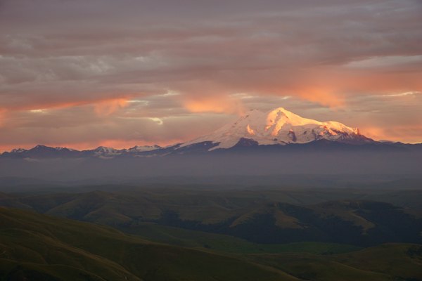 Elbrus glowing at sunset above the plain — Red and white house on green grass field near mountains under white clouds and blue sky