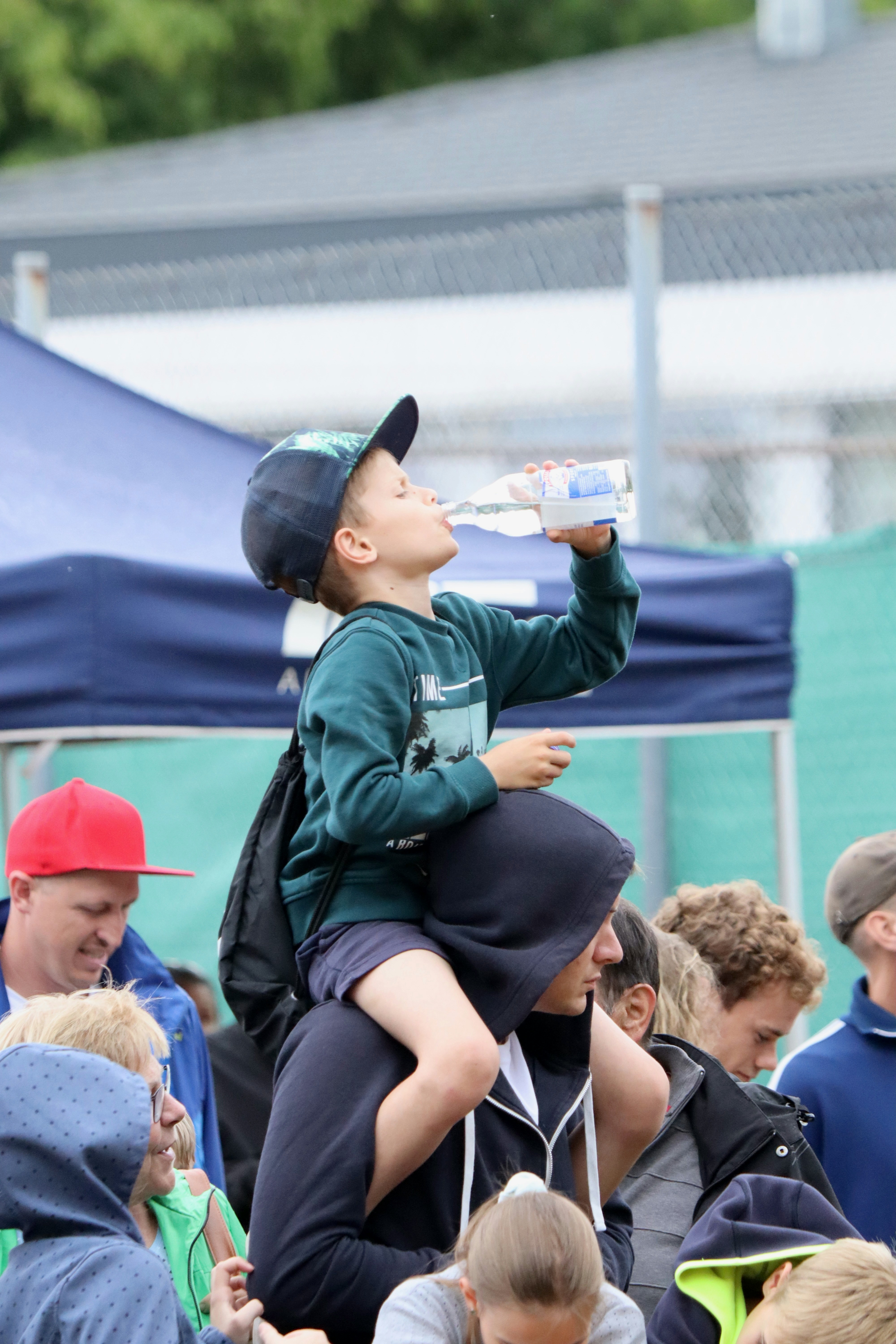 A young boy drinking water from a bottle while sitting on a man's shoulders