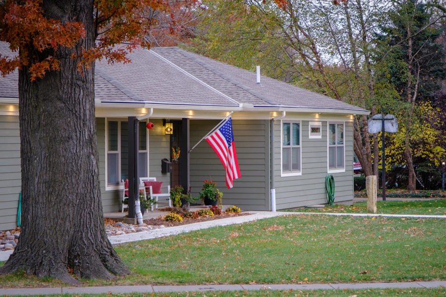 A grey bungalow with a flag on the porch under a big tree