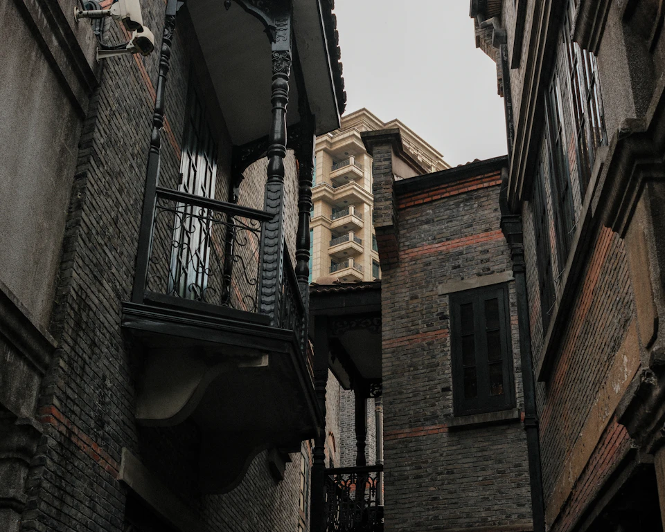 The image depicts a narrow alleyway flanked by two buildings, one with a black balcony and the other with a red brick facade. The building on the left has a black balcony supported by iron bars, while the building on the right features a red brick exterior. Both buildings have multiple windows that reflect the overcast sky above.