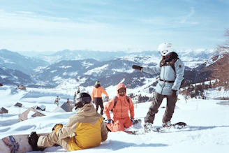 A group of people standing and sitting on top of a snow covered slope
