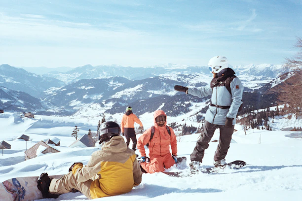 A group of people standing and sitting on top of a snow covered slope